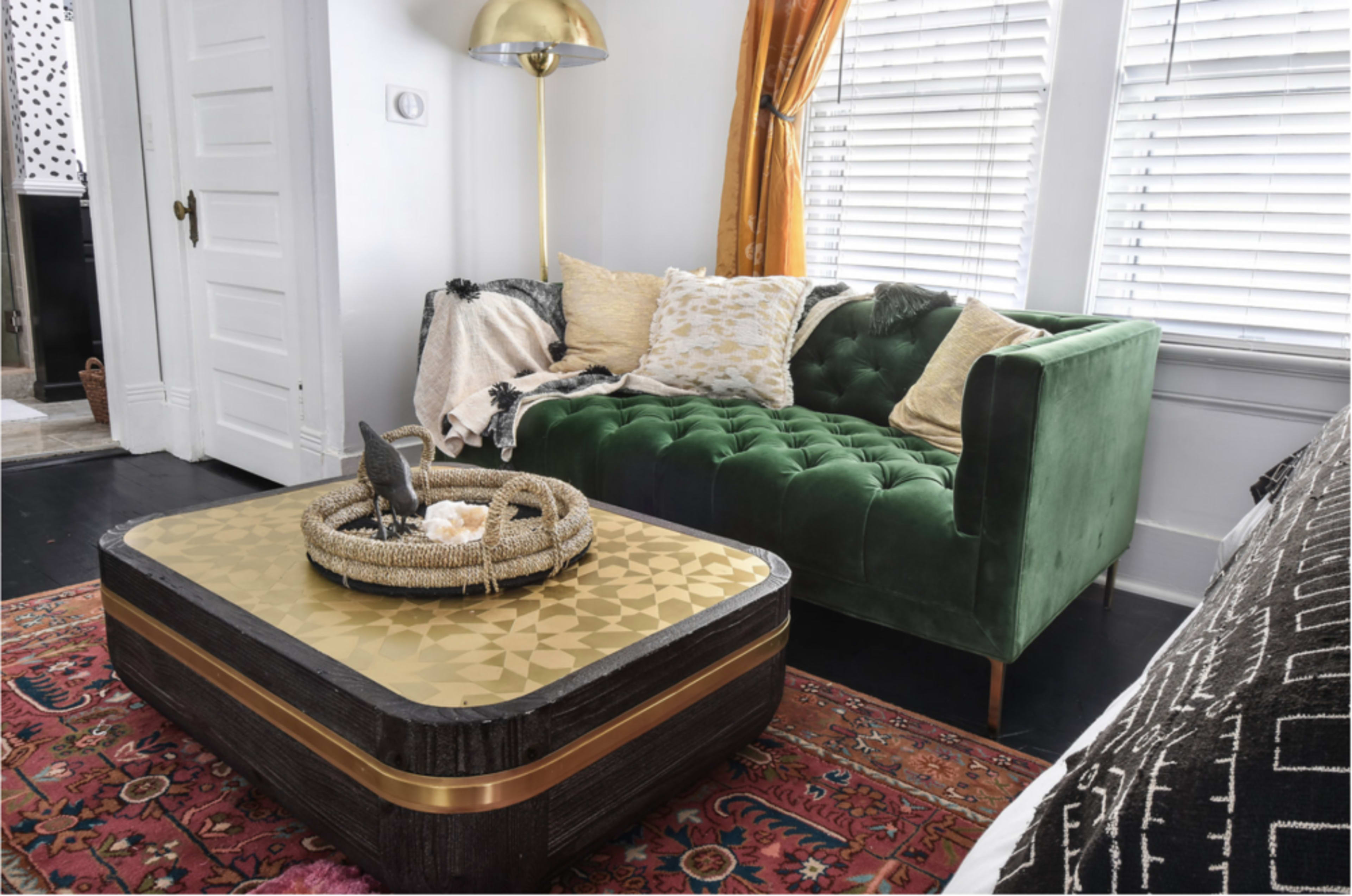 The image shows a green tufted sofa with decorative pillows next to a rectangular coffee table adorned with a tray and a small decorative bird, all set against a backdrop of white walls and large windows.