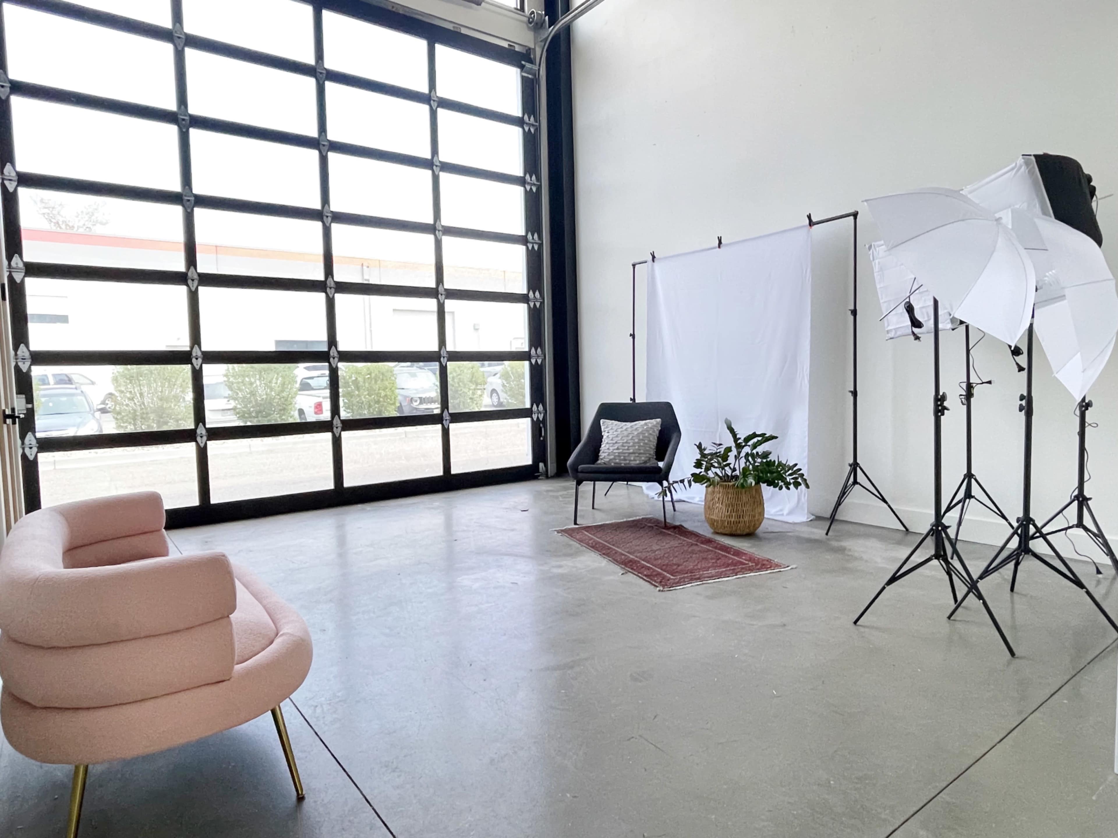 A spacious studio features a pink sofa, a white backdrop supported by stands, and two lighting setups next to a large glass garage door.