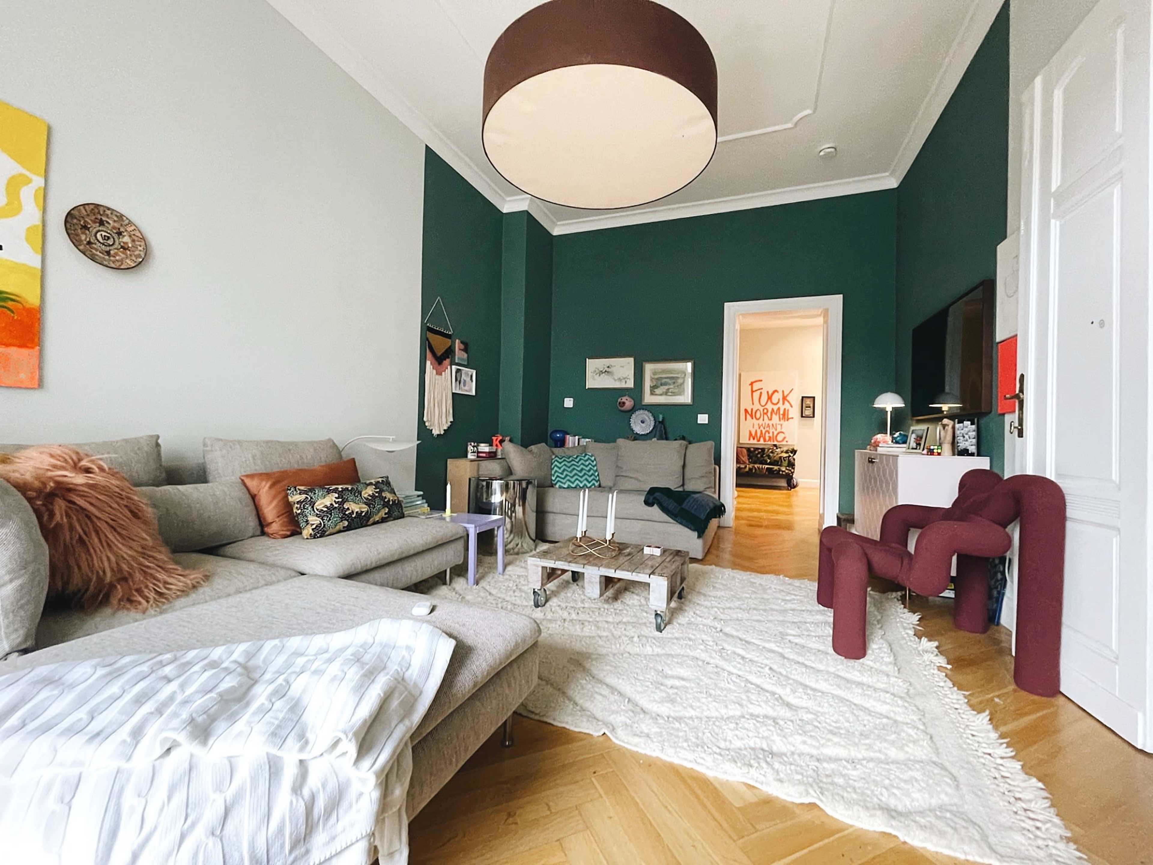 A modern living room features a gray sectional sofa, a maroon accent chair, and colorful wall art against dark green walls.