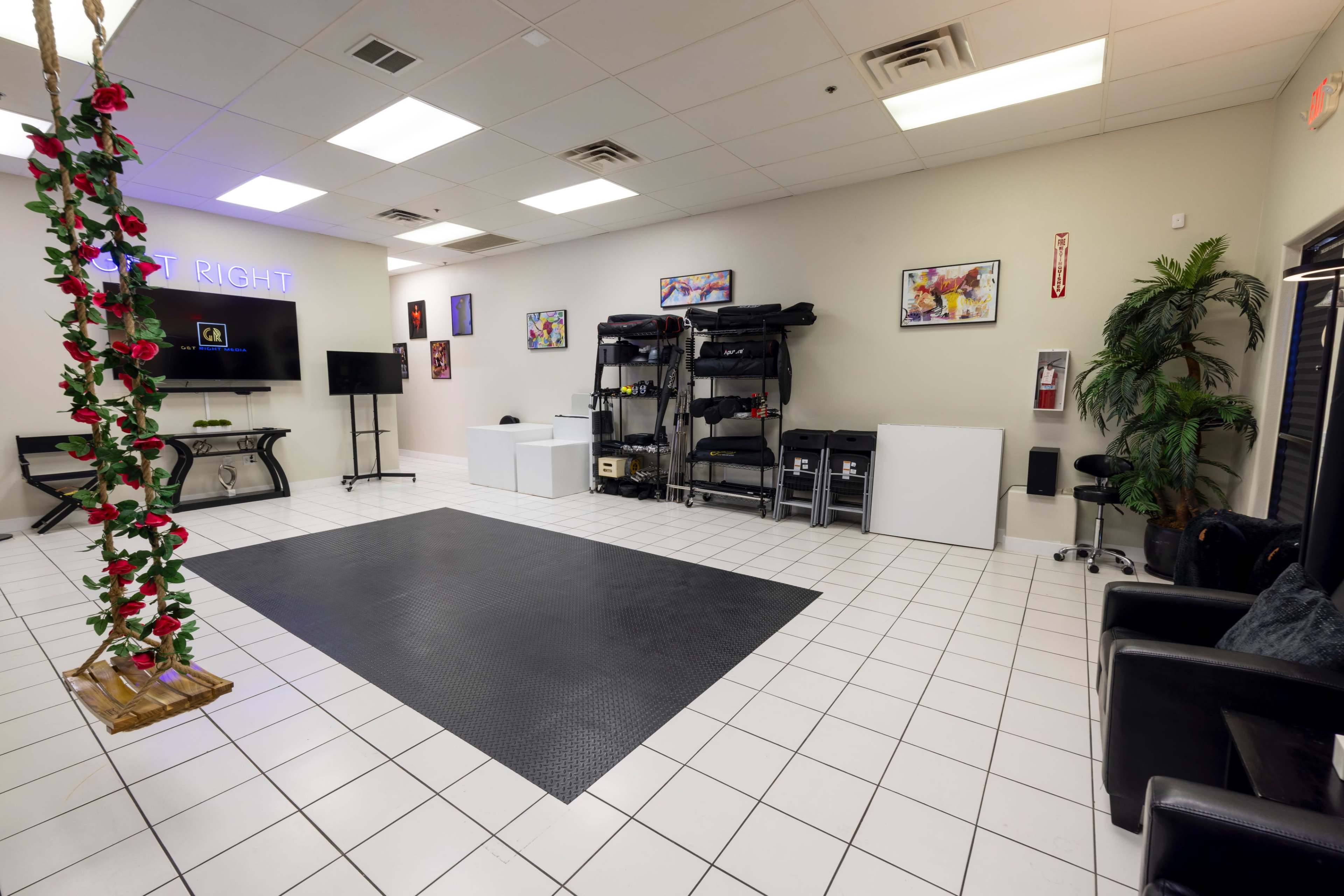The image shows a spacious room with a black rubber mat, wall-mounted shelves filled with equipment, and a swing decorated with artificial flowers hanging from the ceiling.