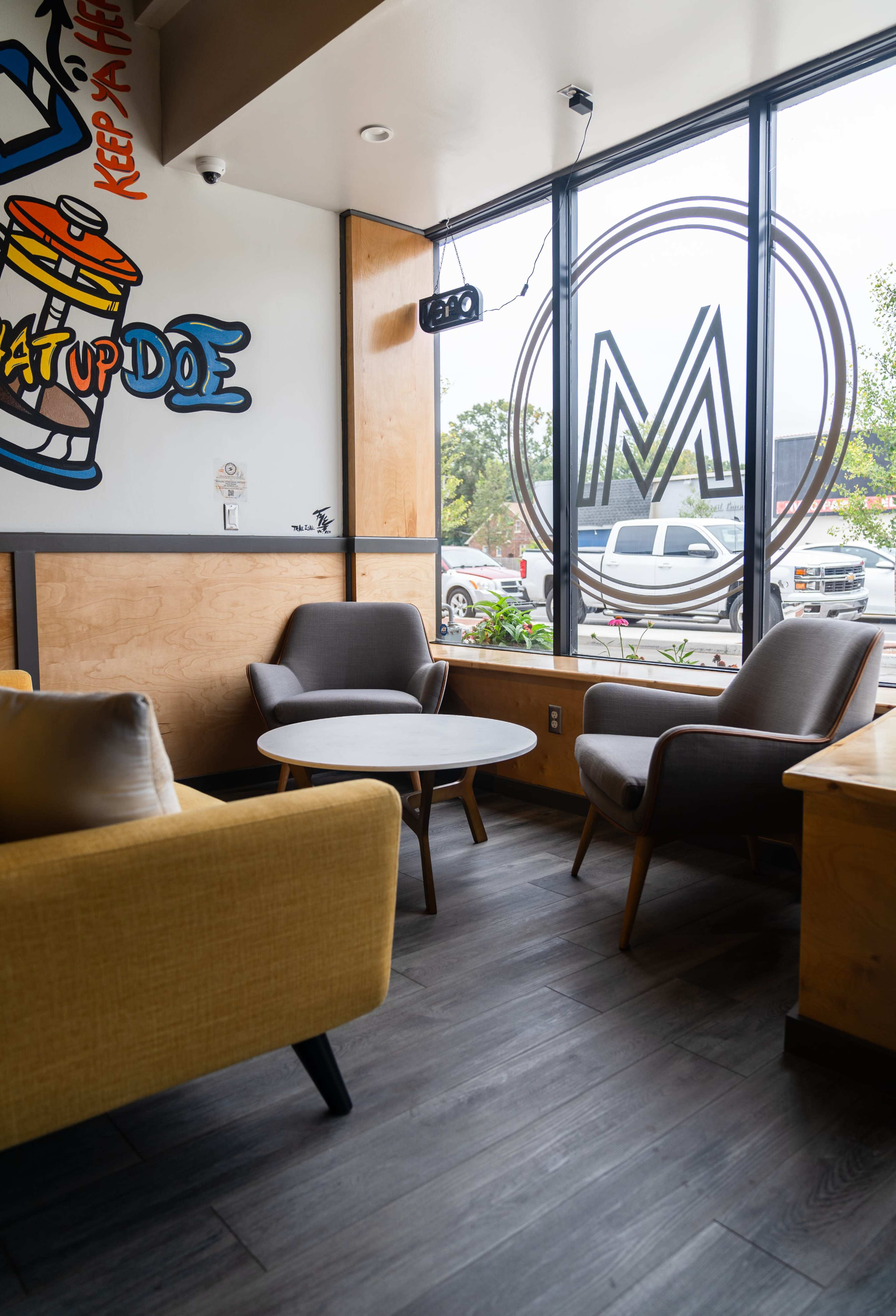 A cozy café interior with two gray armchairs, a round table, and large windows displaying a view of an outdoor scene.