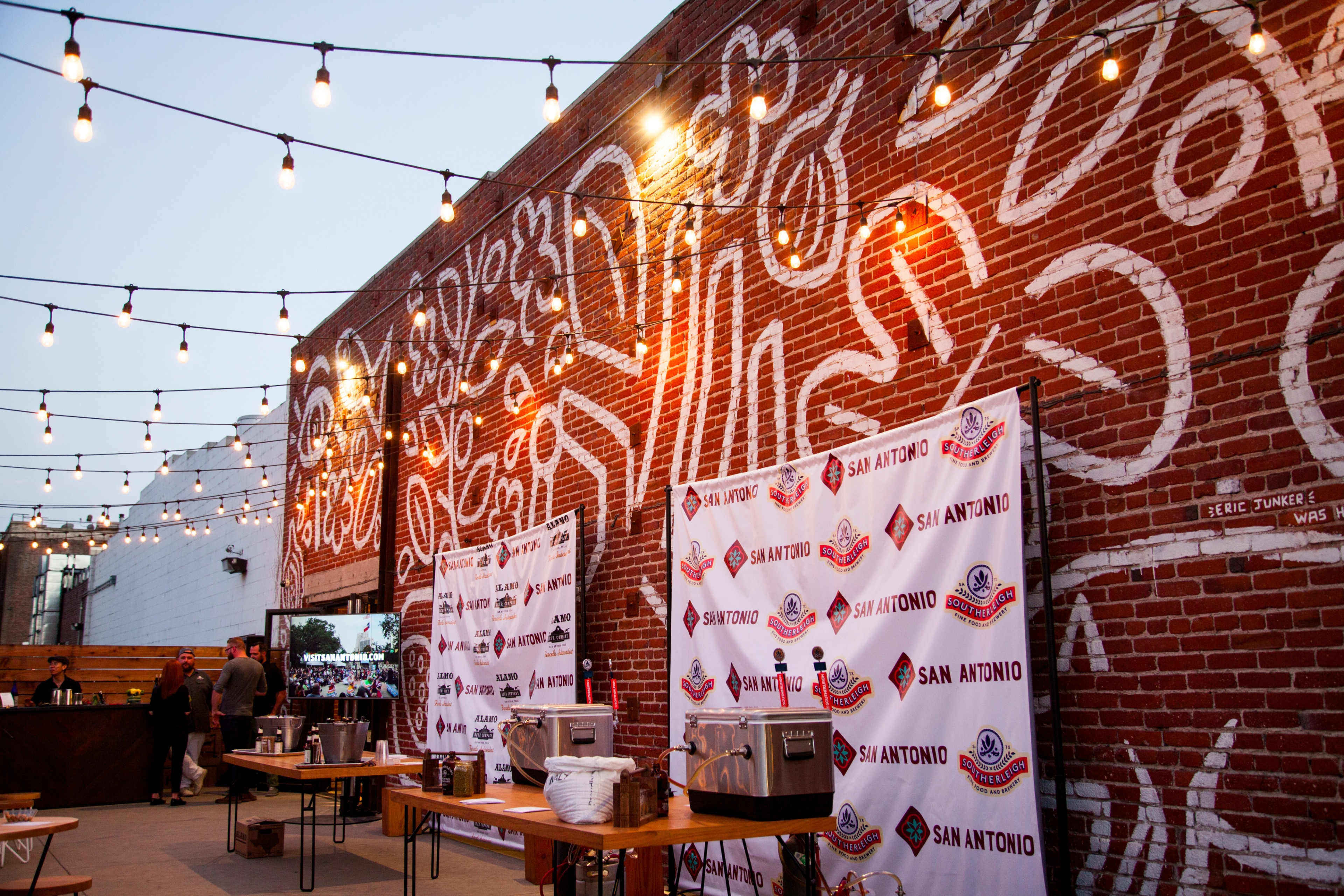 An outdoor event space with string lights above, featuring large murals on a brick wall and tables set up with catering equipment.