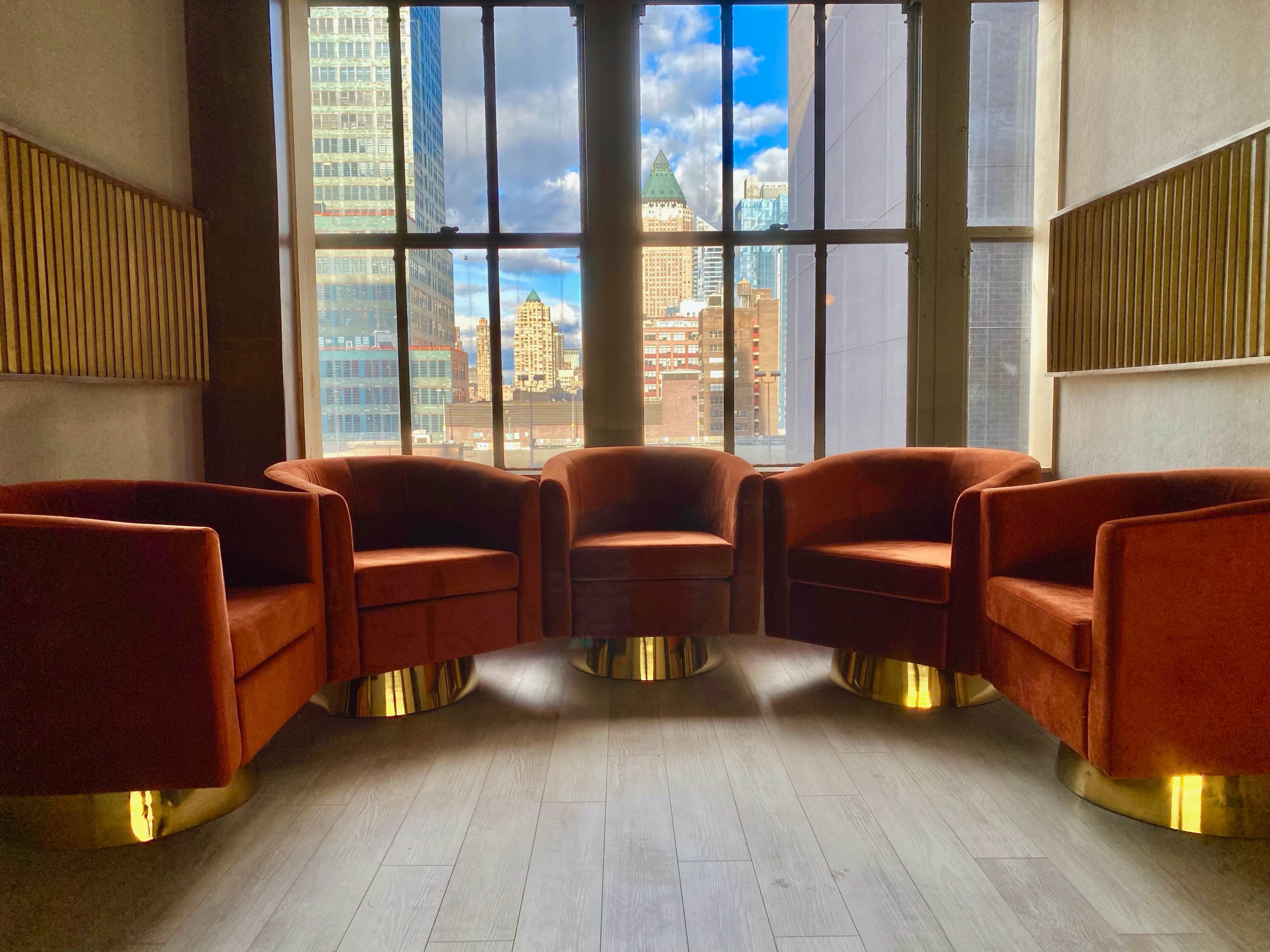 Four orange velvet chairs with gold bases are arranged in a circle near large windows, overlooking a city skyline.