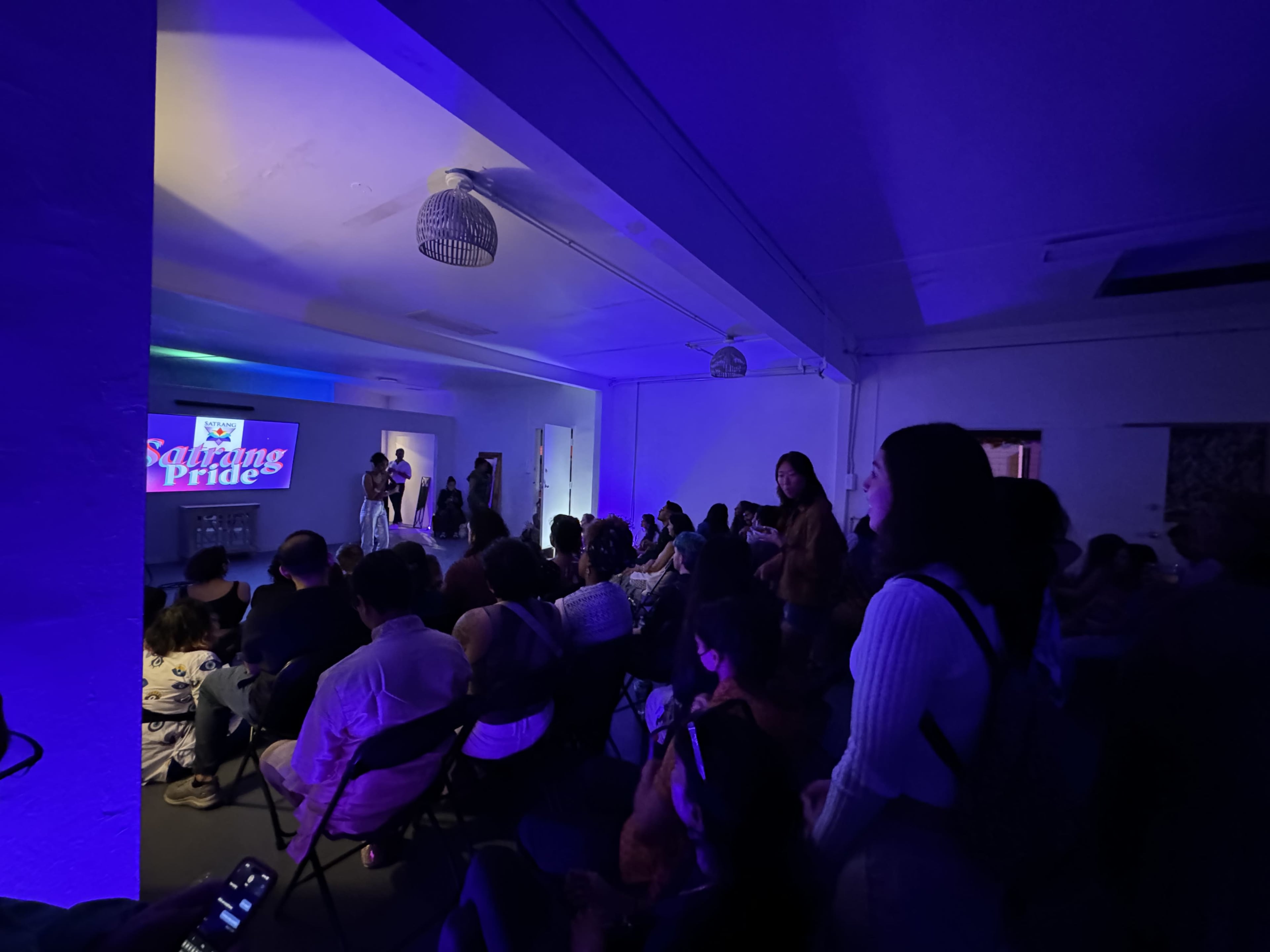 A diverse group of people is seated in a dimly lit room, watching a presentation titled "Sarang Pride" on a large screen.
