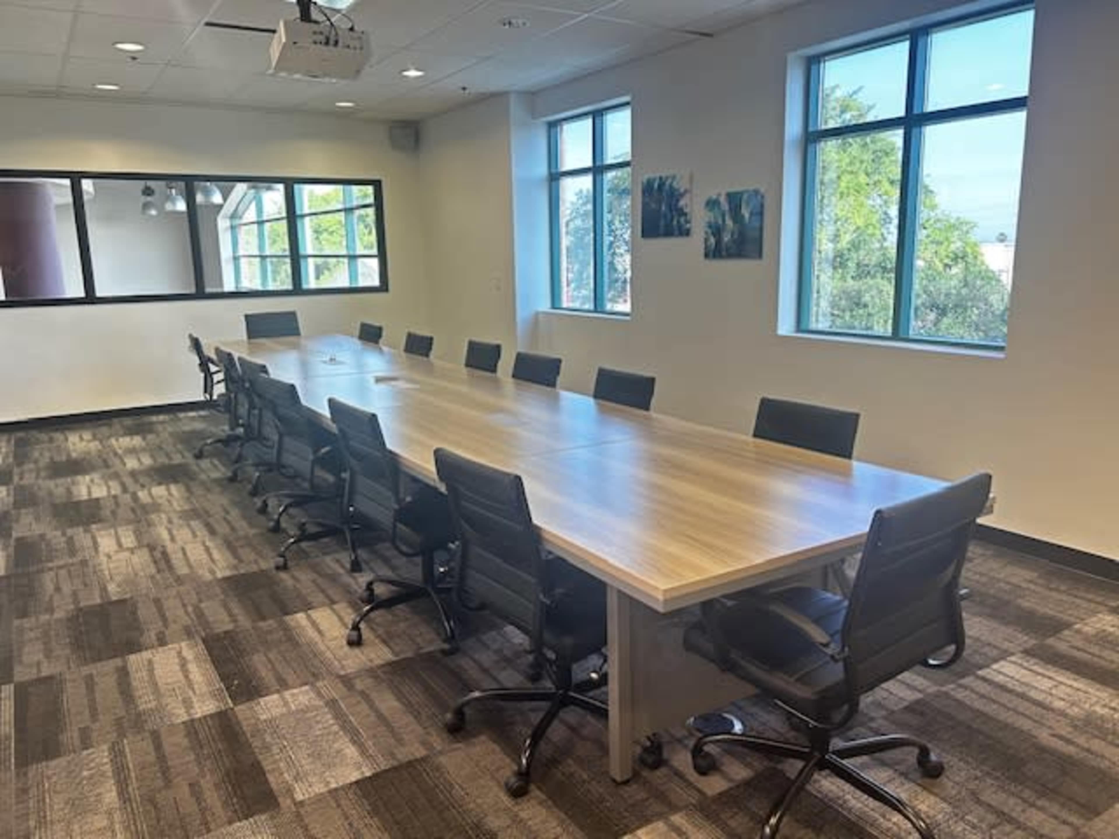 A long conference table surrounded by black office chairs is displayed in a well-lit room with windows and wall-mounted artwork.