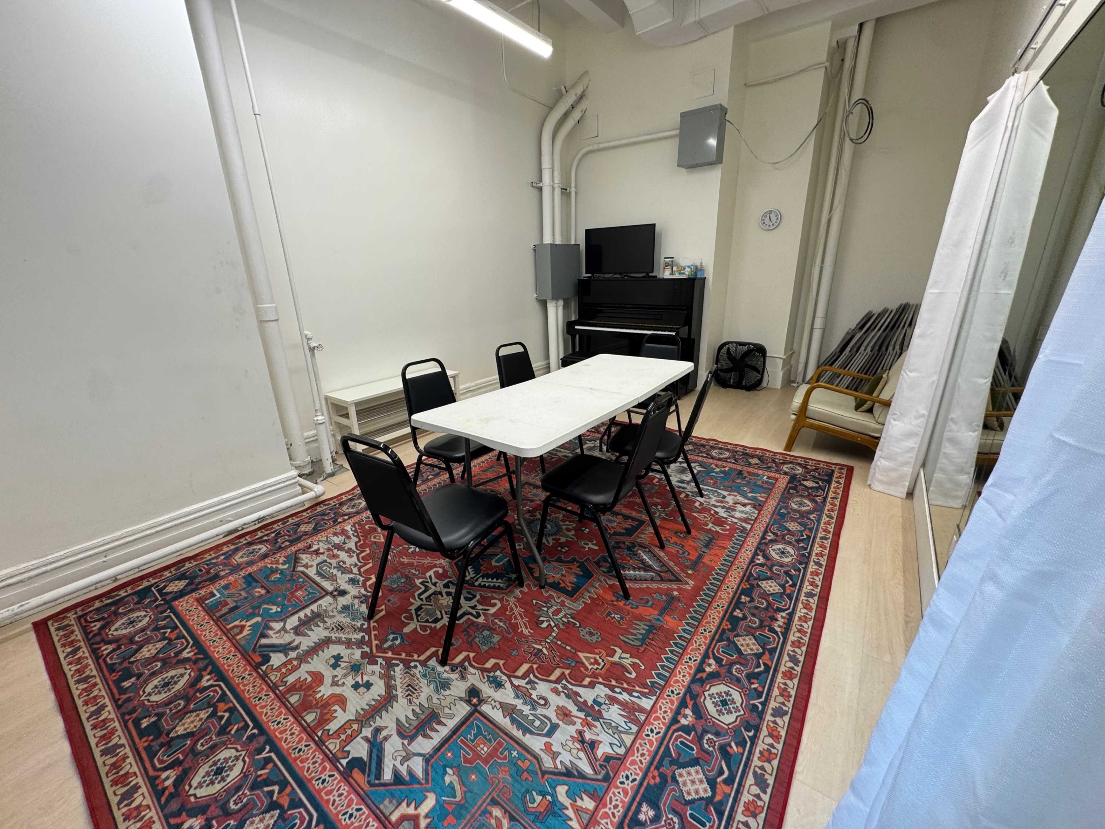 The image shows a small room with a rectangular table and five black chairs arranged on a large patterned rug, featuring a piano in the corner and a mirror along one wall.