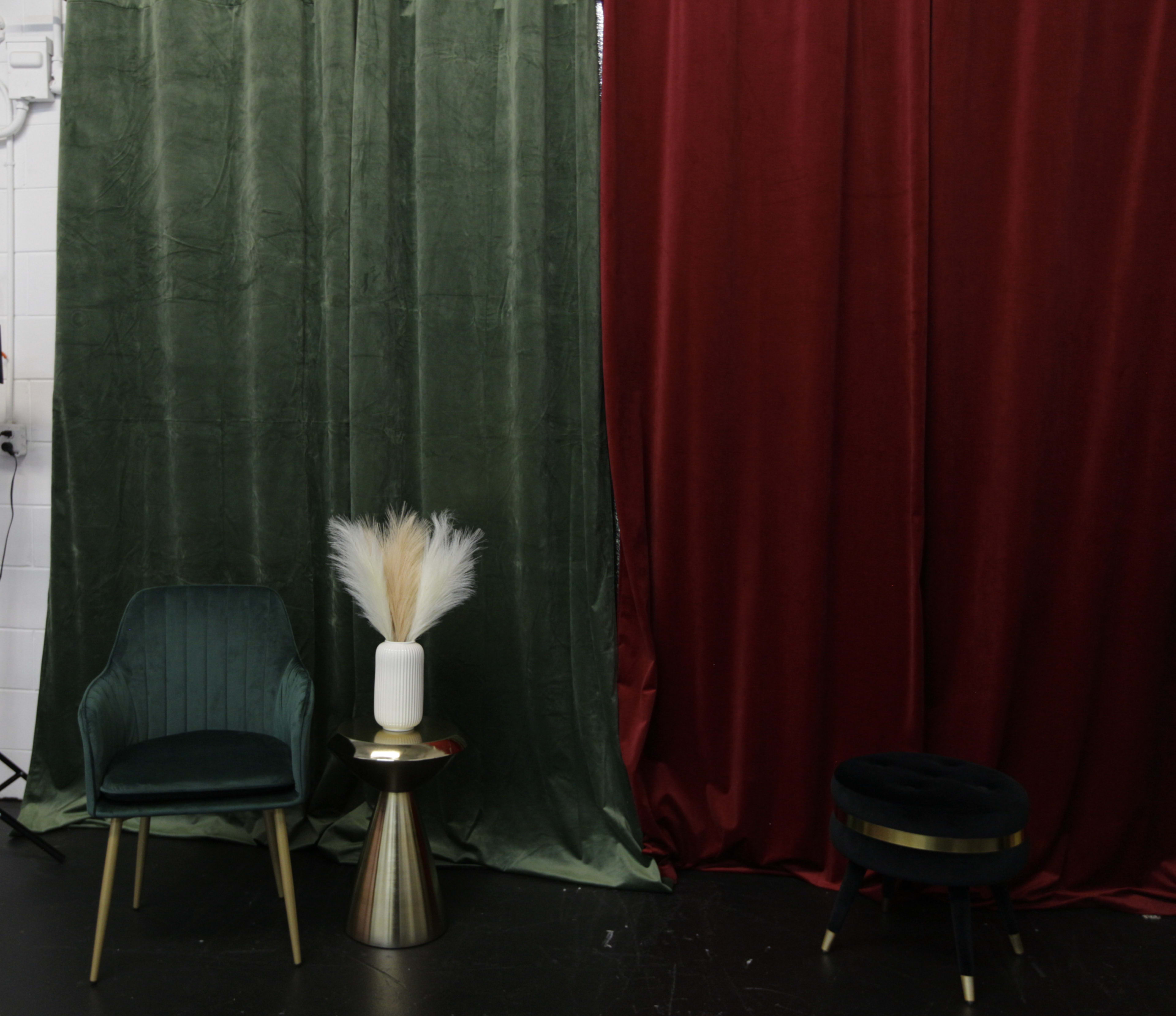 A green velvet curtain and a deep red curtain frame a small, styled corner featuring a chair, a metallic side table with a vase, and a round ottoman.