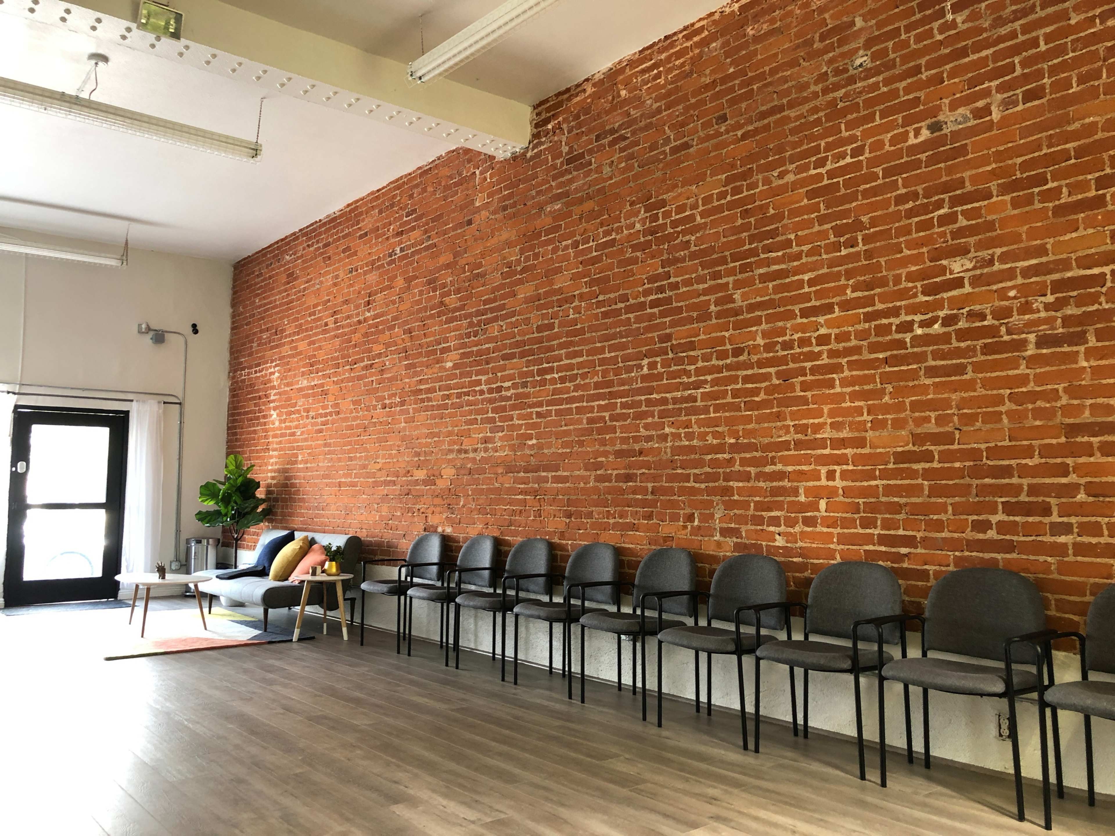 A waiting area with a row of gray chairs, a couch, and a brick wall in a well-lit space.
