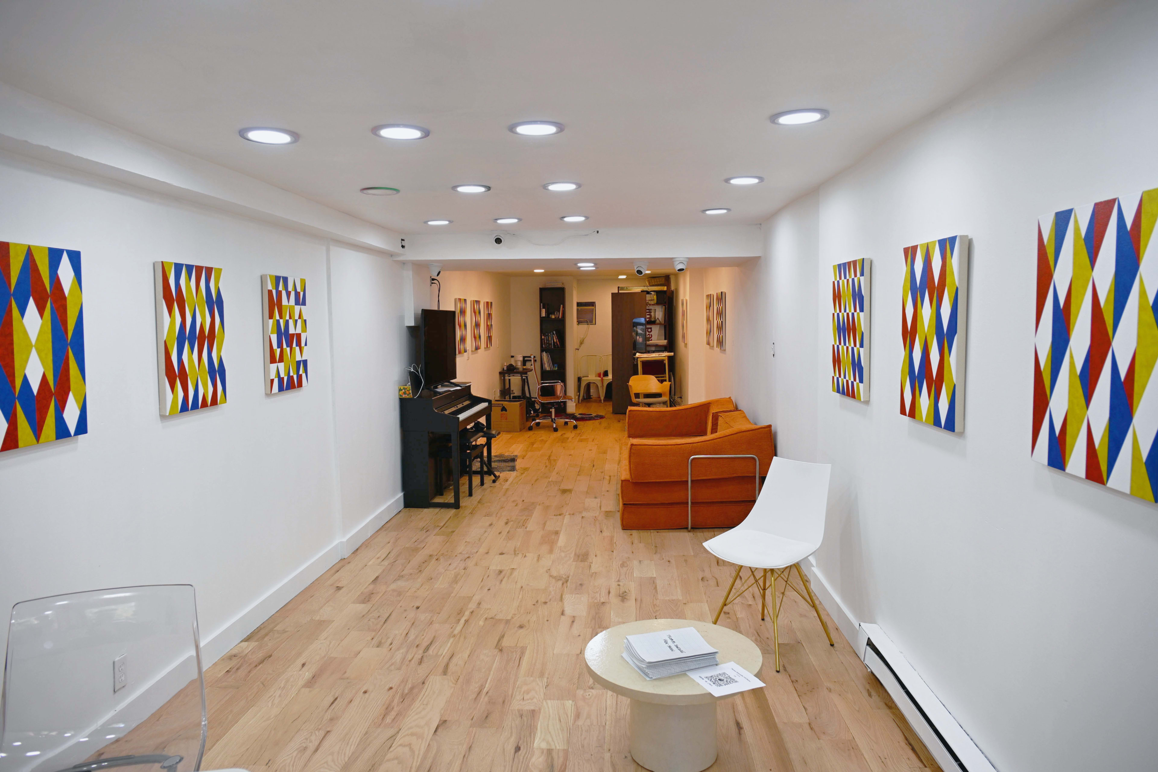 The image shows a well-lit gallery space with wooden flooring, featuring colorful geometric artwork on white walls, a piano on one side, and orange seating along with a white chair.