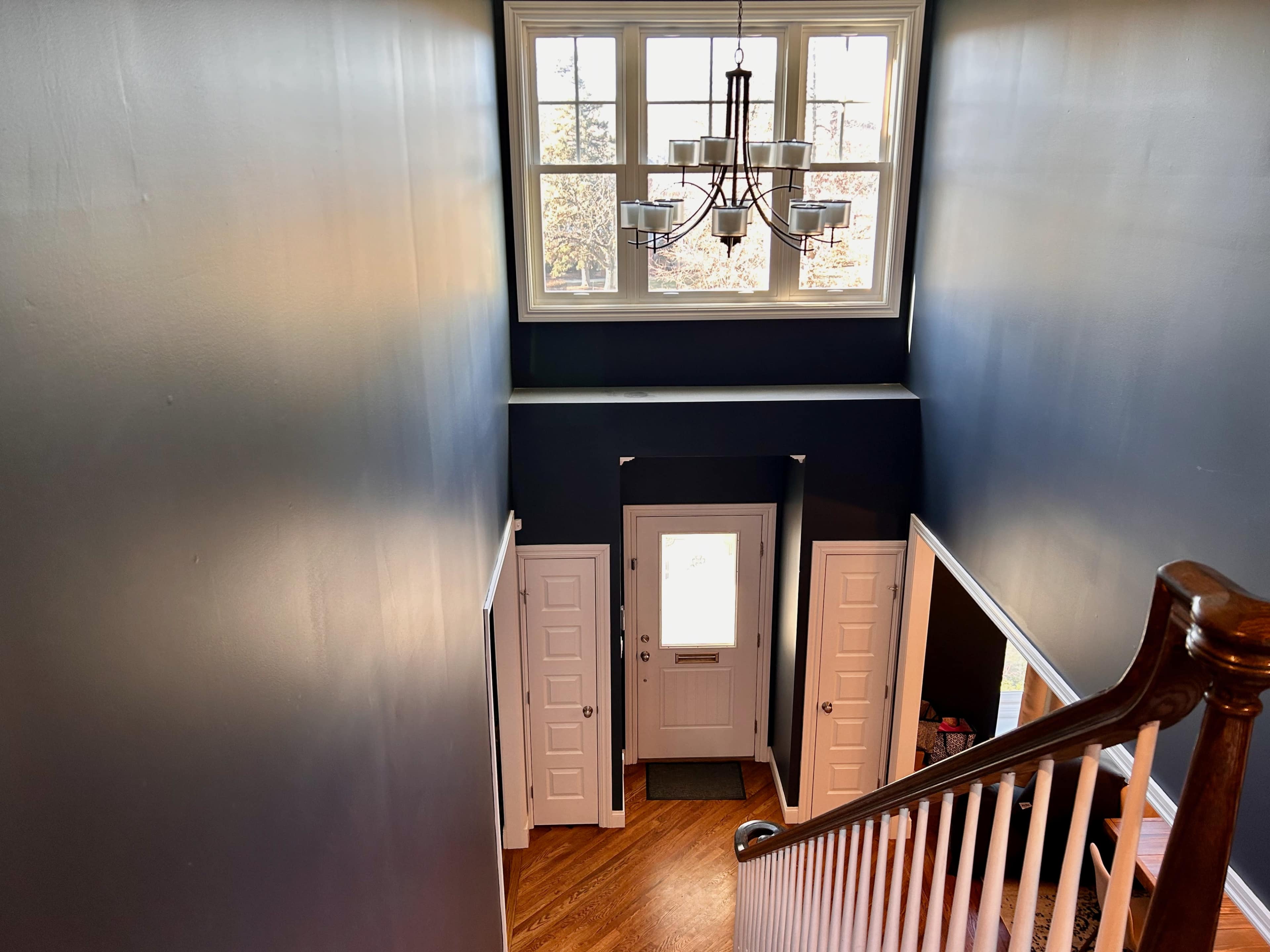 A staircase descends from a second floor to a front door, flanked by white doors and illuminated by a chandelier and natural light from a large window above.