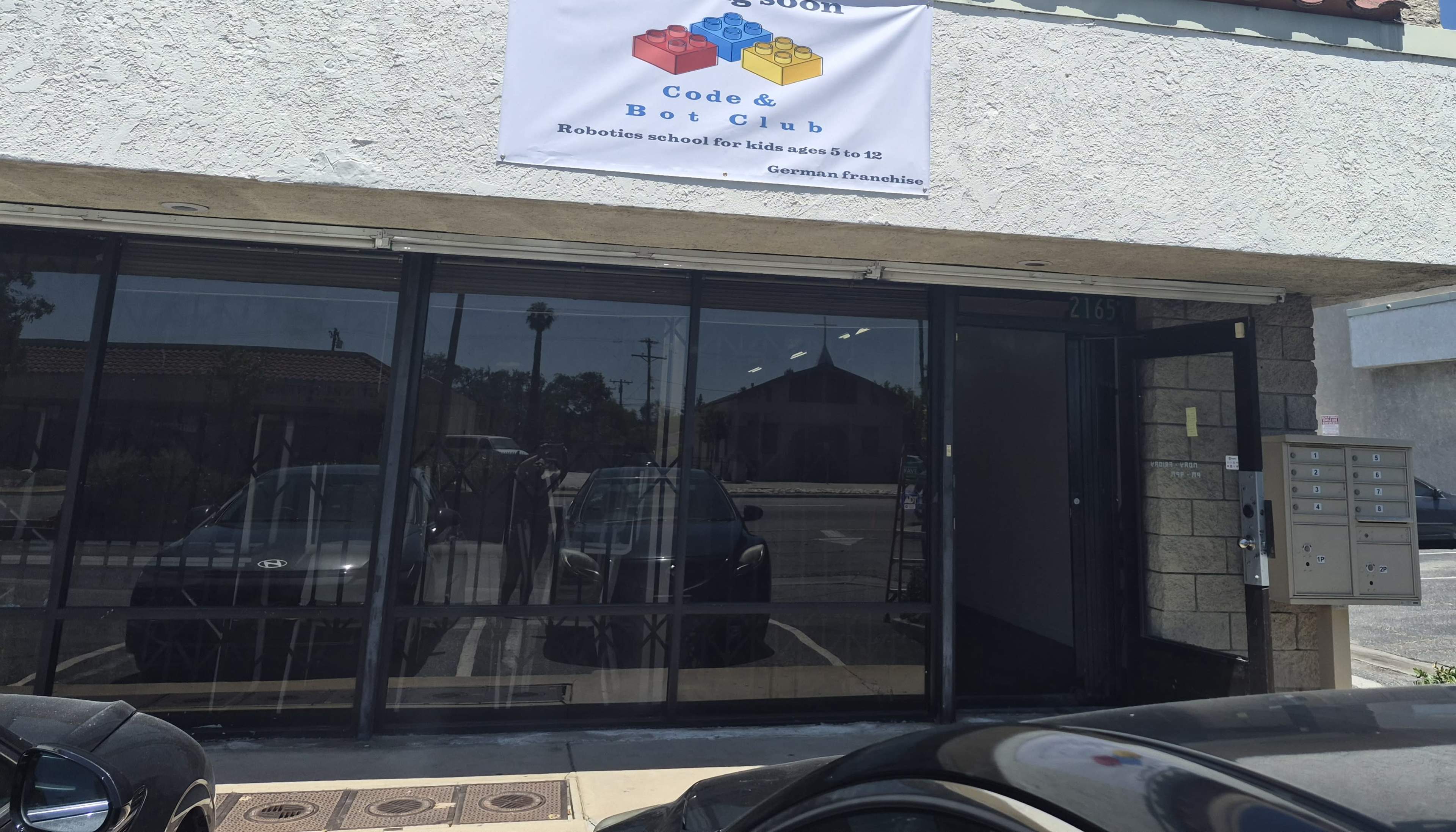 The image shows the exterior of a building with a sign for "Code & Bot Club," a robotics school for kids, and features a glass door with visible mailboxes nearby.