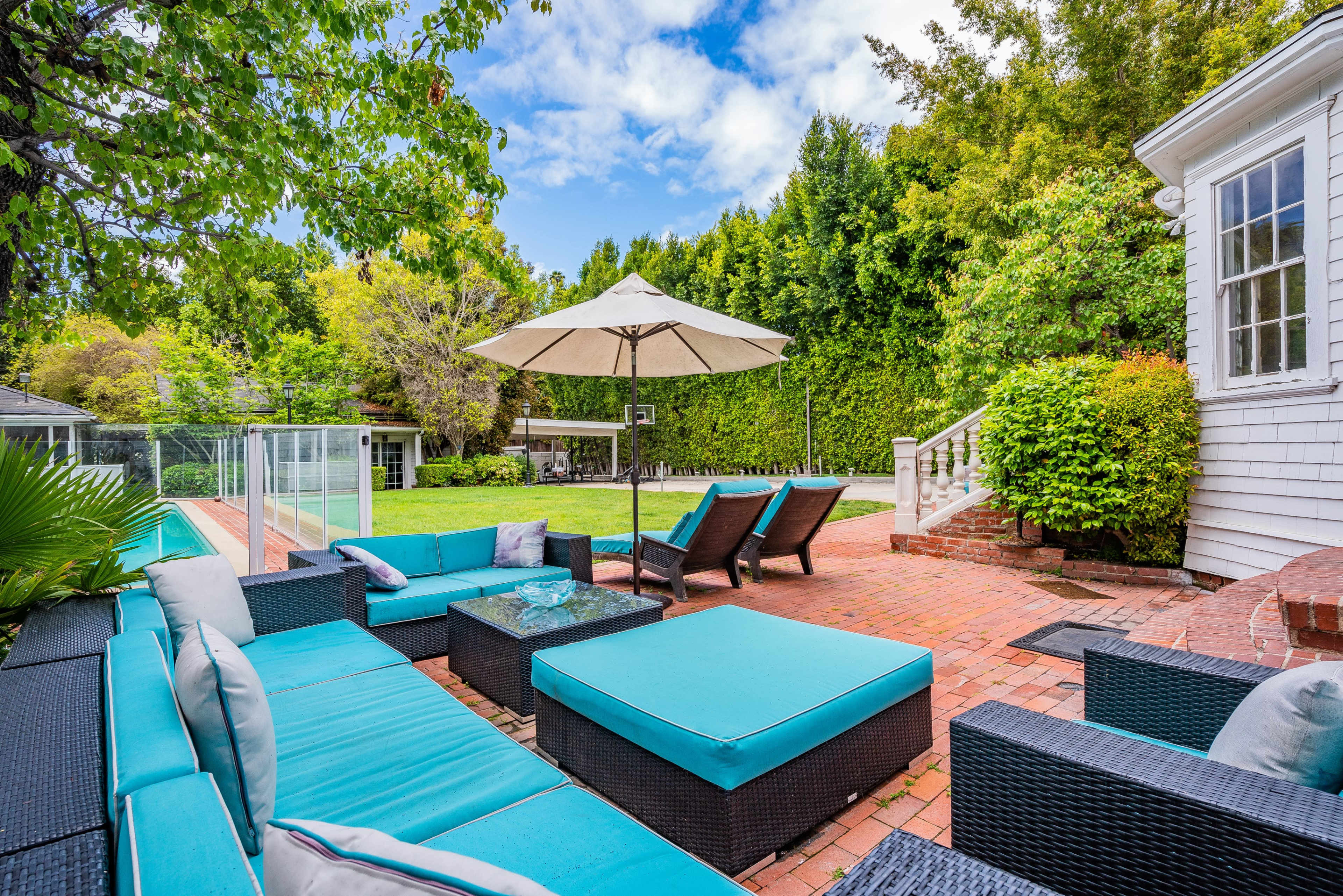 A backyard patio area with turquoise lounge furniture, an umbrella, and a swimming pool surrounded by greenery.