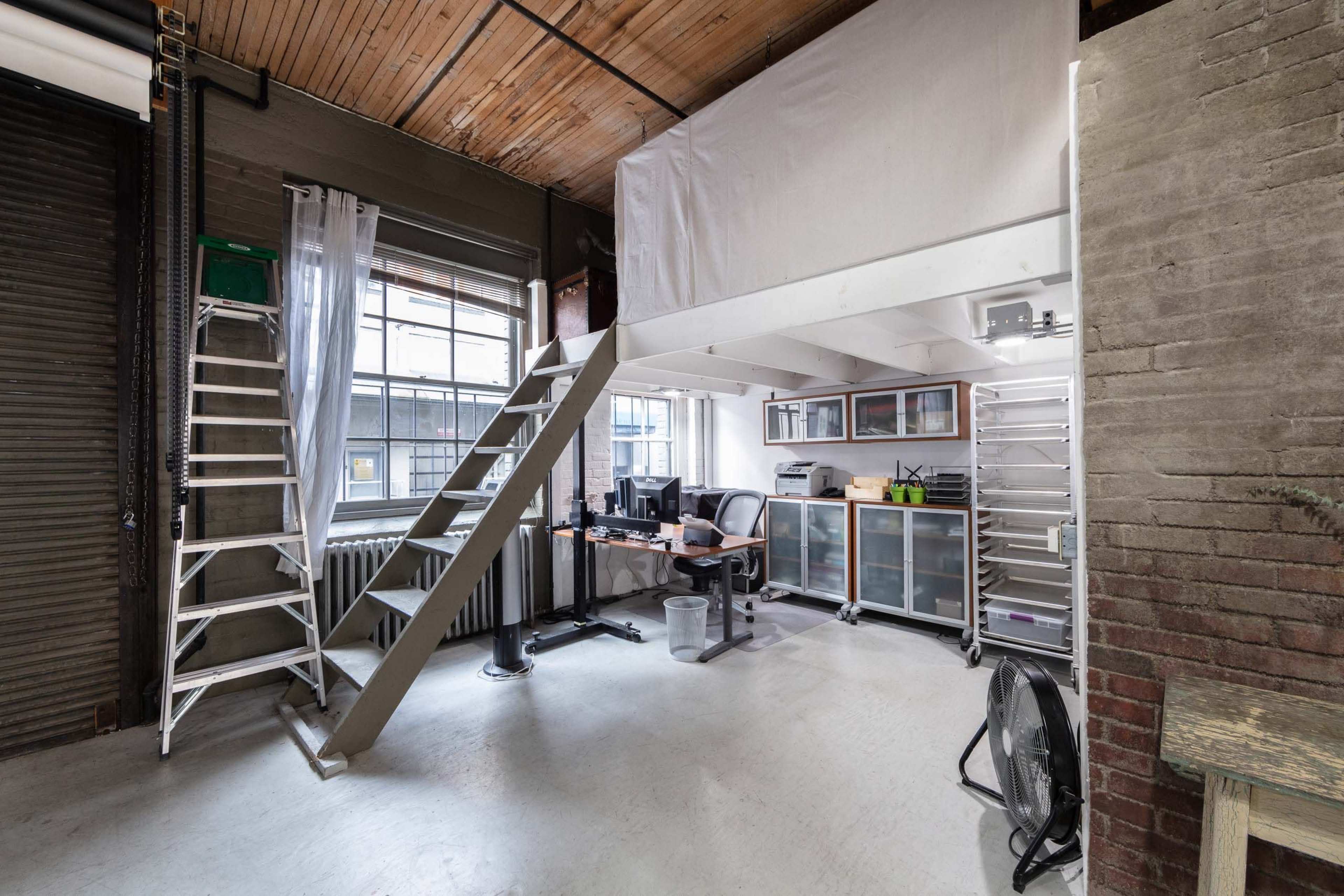 A modern office space with a lofted area, featuring a desk with computers, storage units, and a ladder against a wall.