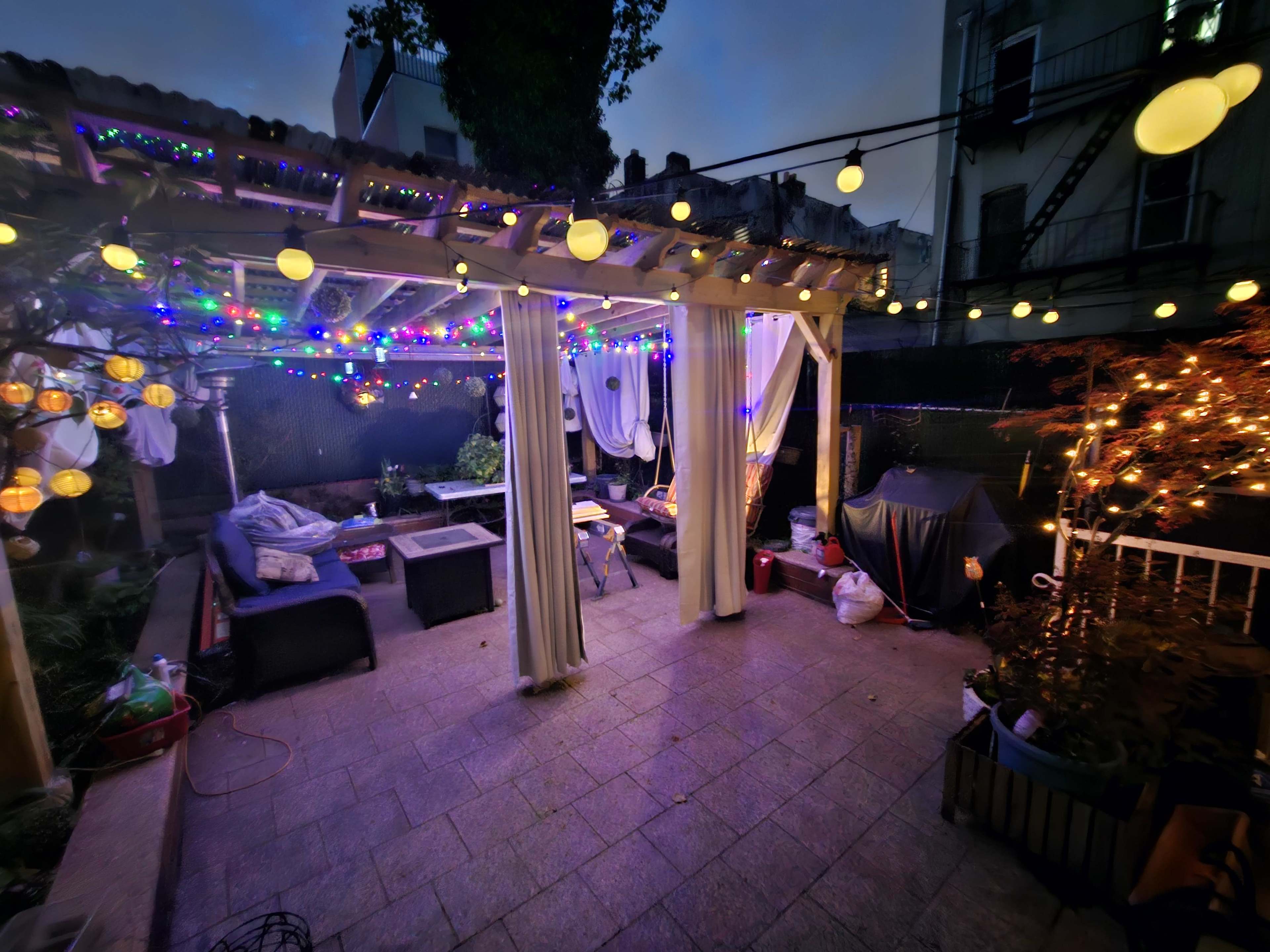 A dimly lit outdoor patio with string lights and lanterns, furnished with several seating areas and decorated with plants.