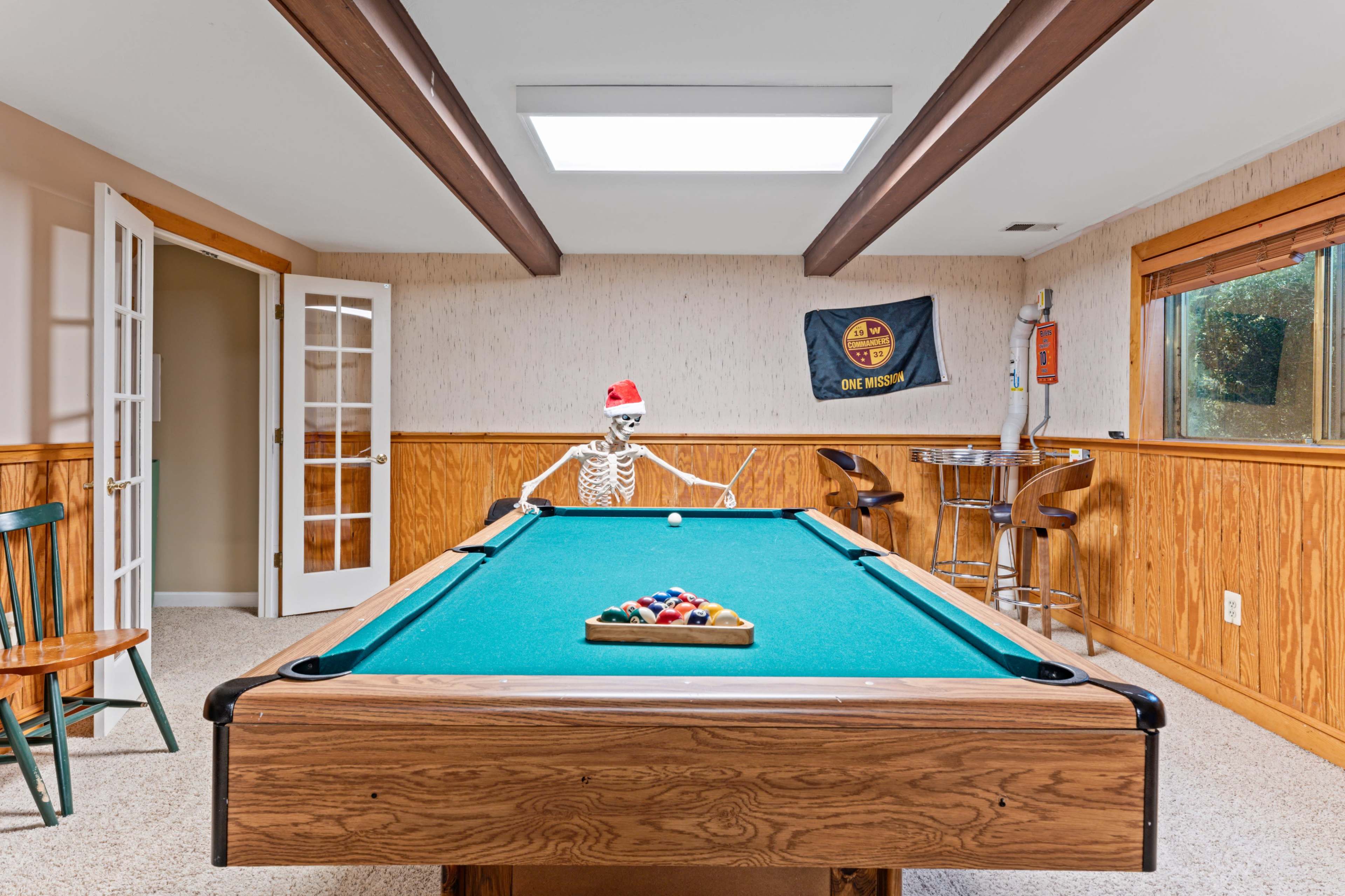 A skeleton wearing a Santa hat sits at a pool table in a rec room with wooden paneling and a bar area in the background.
