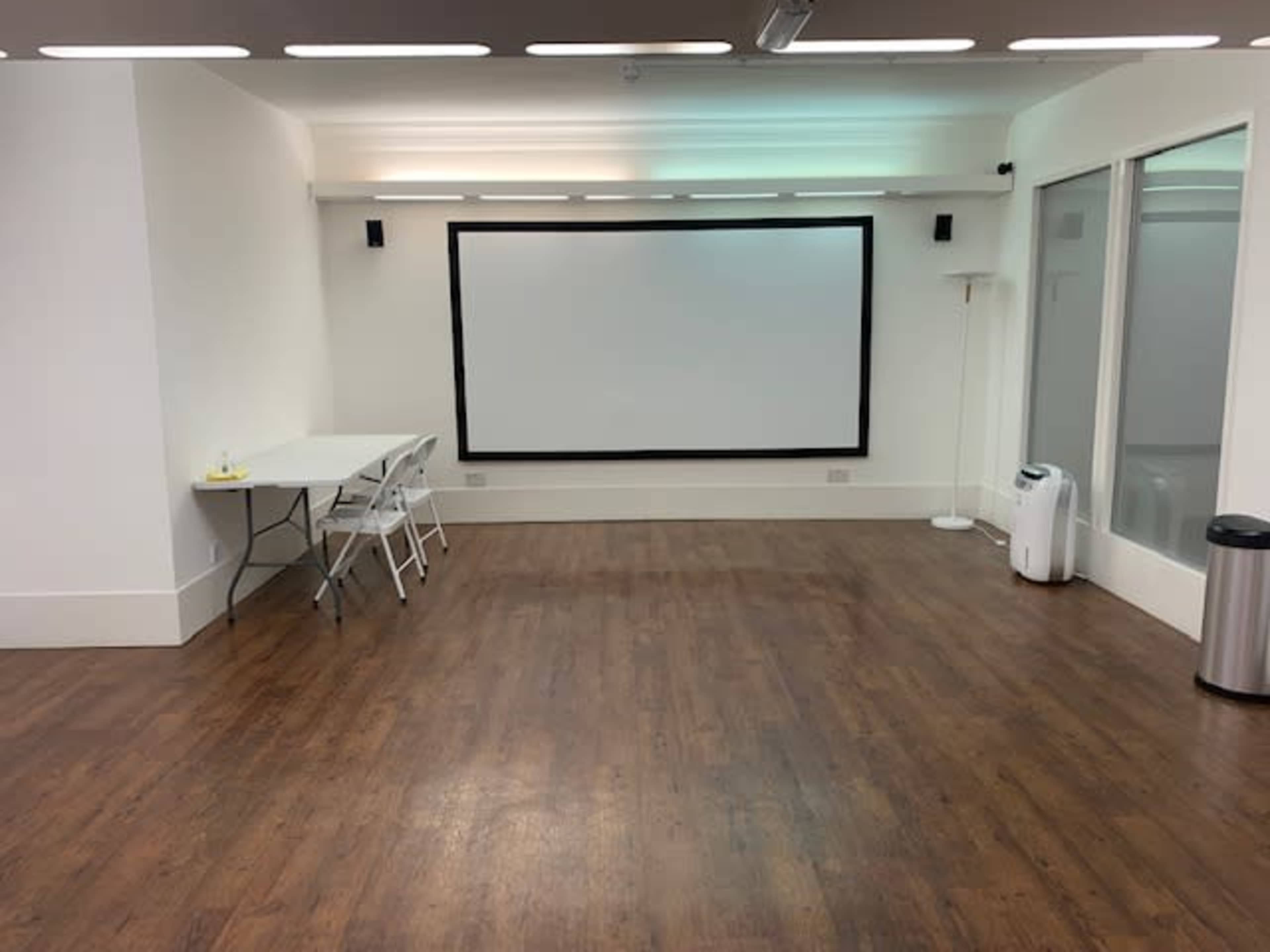 A spacious room with hardwood flooring features a large projection screen, a folding table with chairs, and a trash can in the corner.