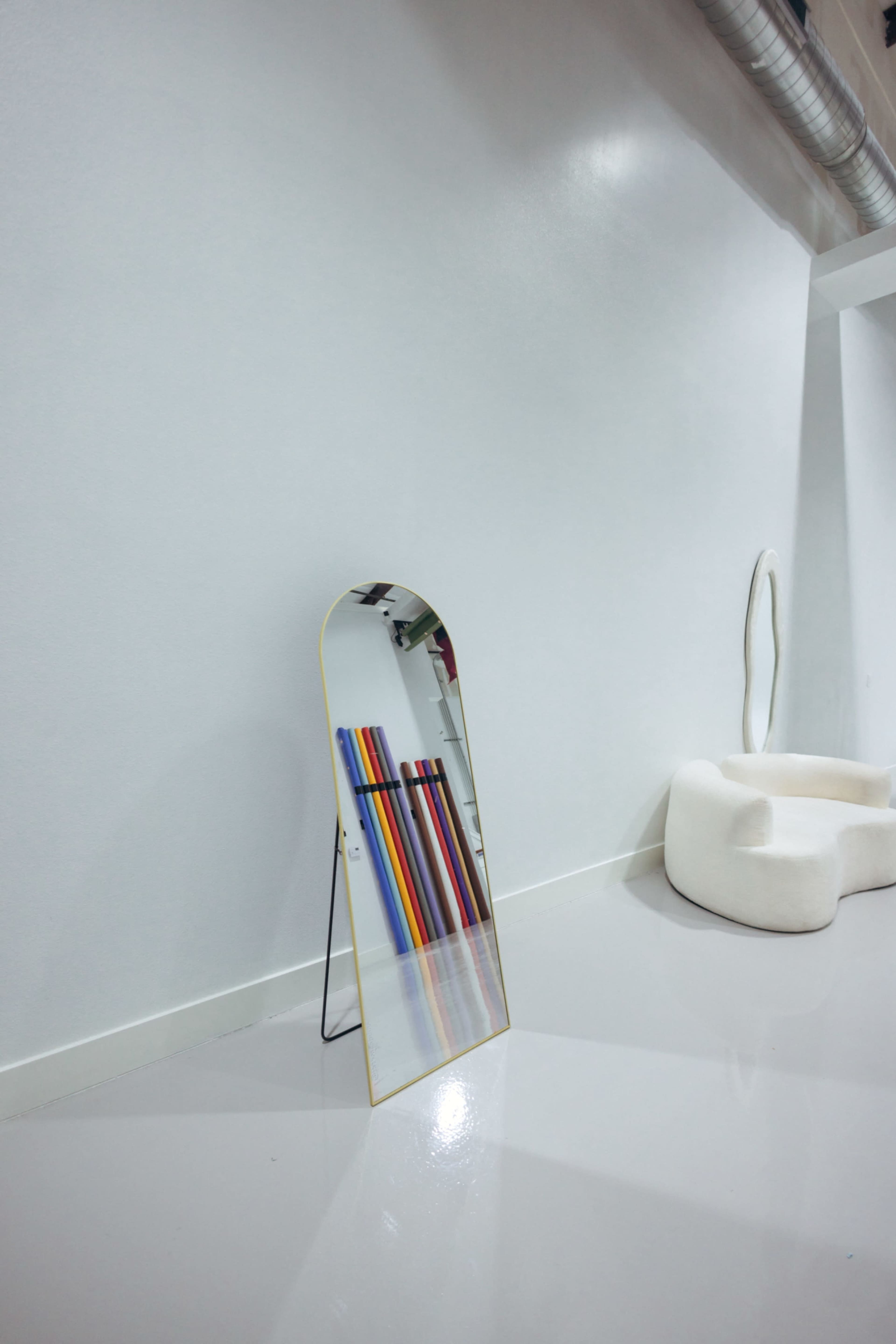A tall, arched mirror stands on a black metal frame against a white wall, beside a white sculptural chair and several colorful poles leaning against the wall.