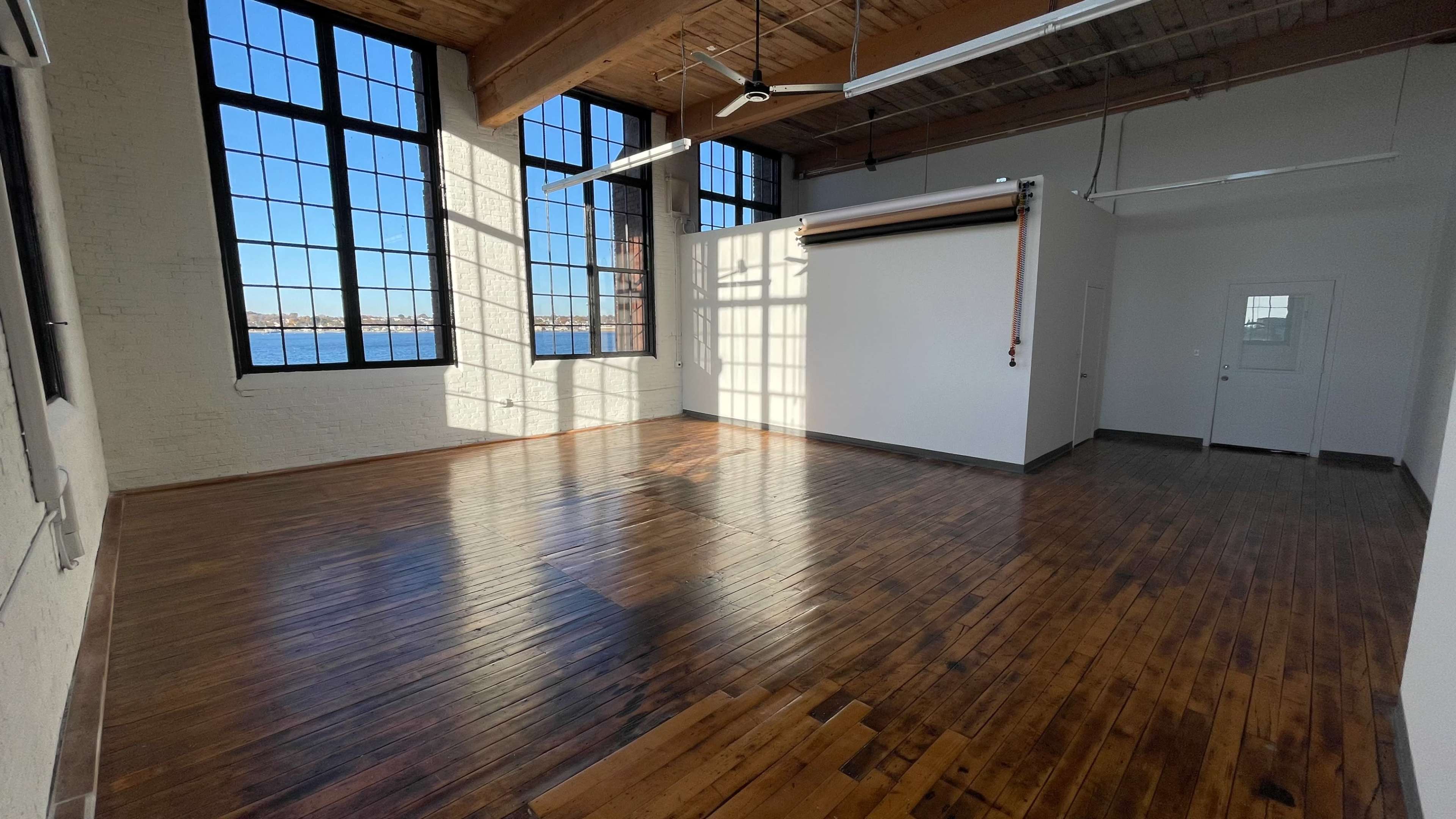 A spacious room with large windows, wooden floors, and a white partitioned wall.
