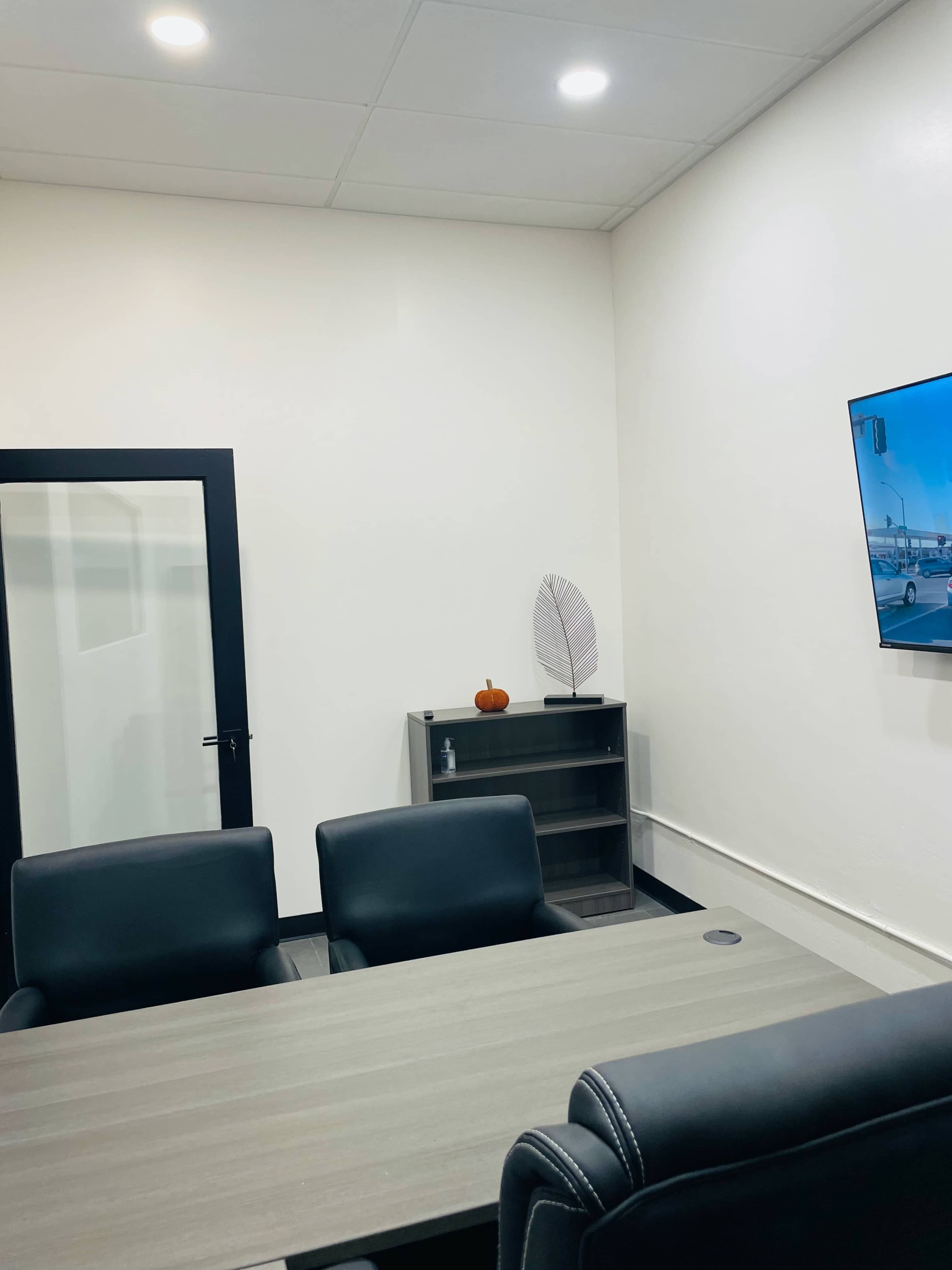 A meeting room features a table with black chairs, a shelf with a decorative item, and a television mounted on the wall.
