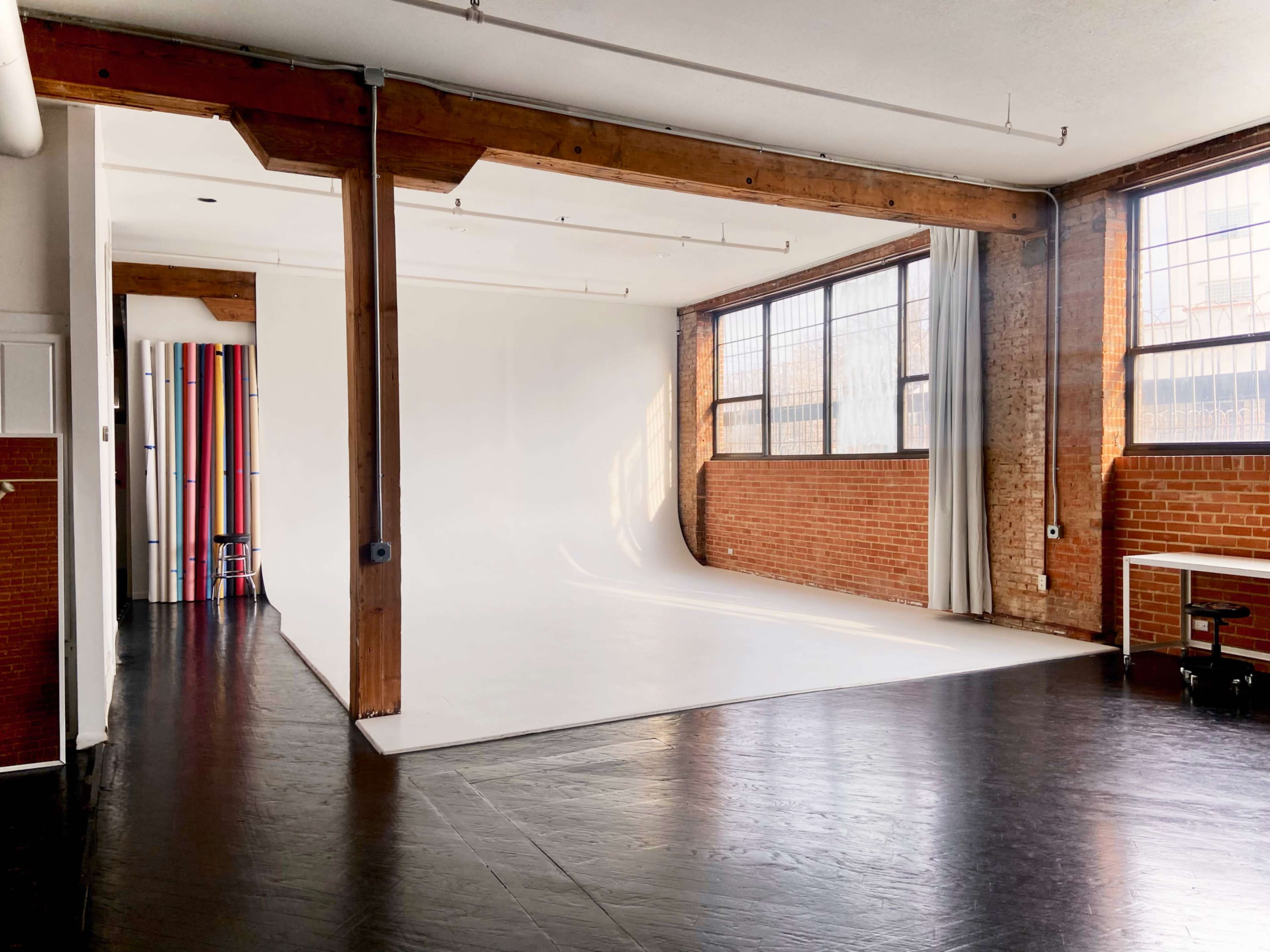 The image shows a spacious photography studio featuring a white backdrop, wooden beams, large windows, and a wooden floor.