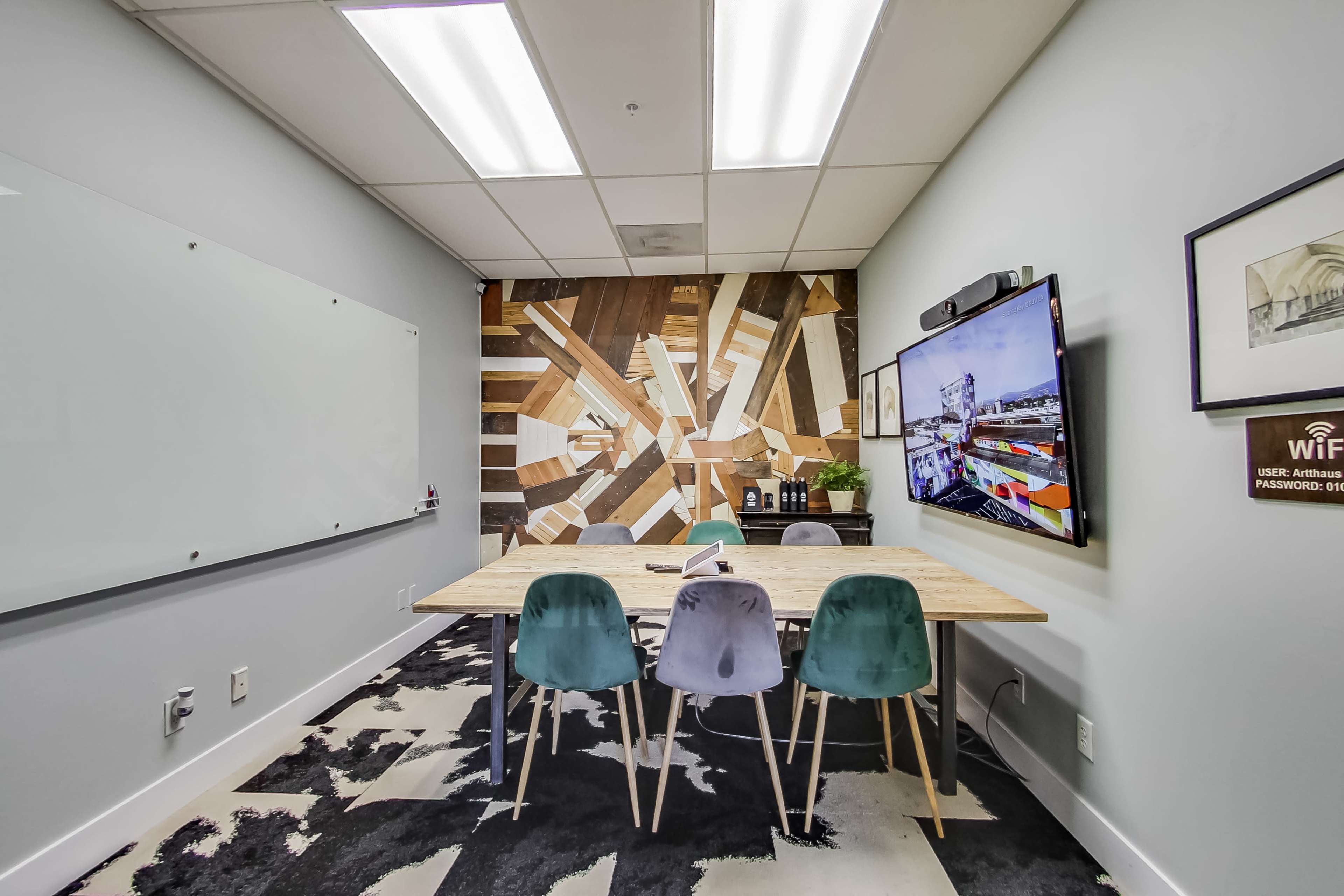 A small conference room features a wooden table surrounded by four chairs, a wall-mounted television, and a large geometric mural behind the table.