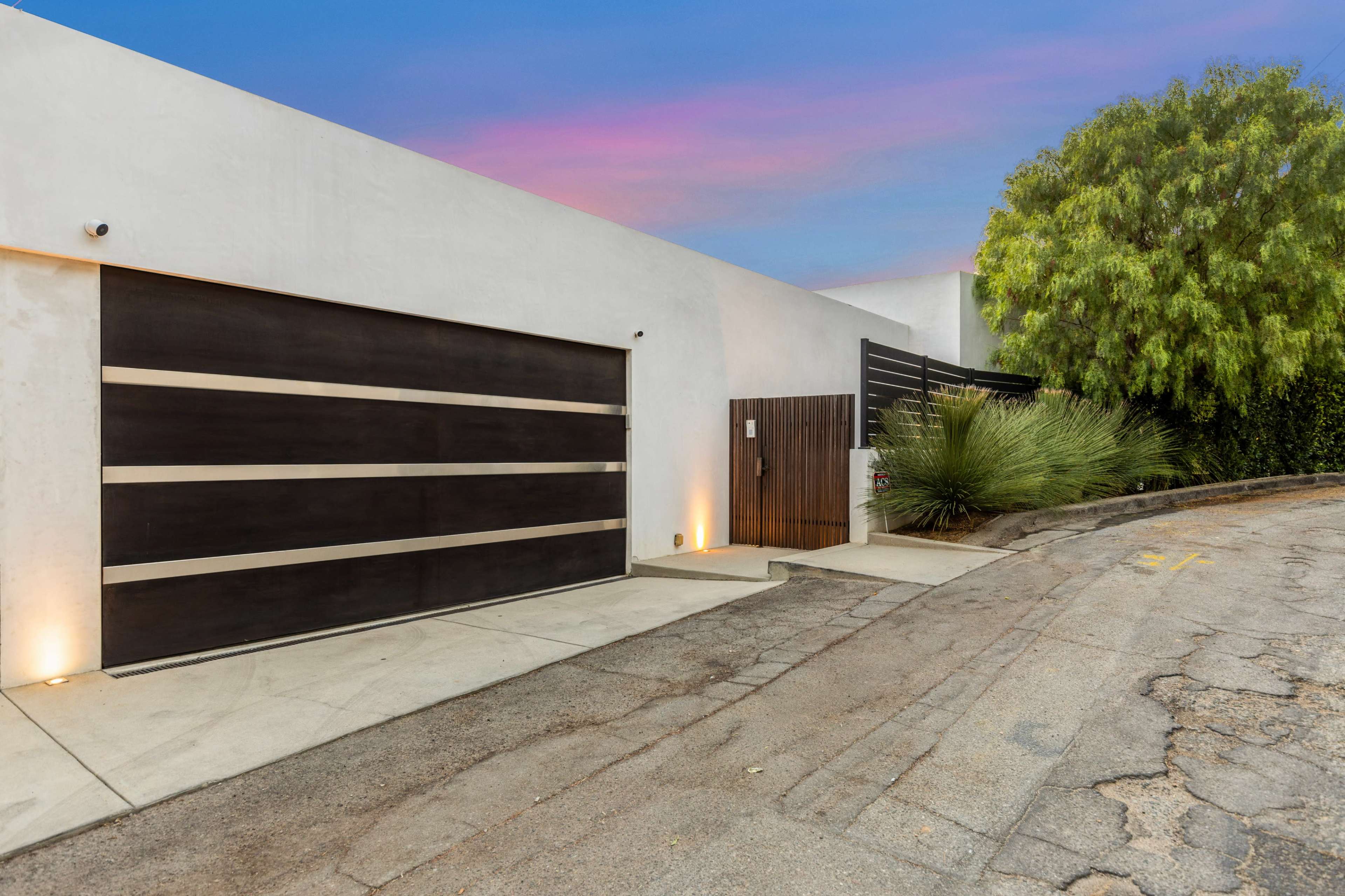 The image shows a modern house with a garage door featuring horizontal wooden slats, situated on a sloped driveway with minimal landscaping.