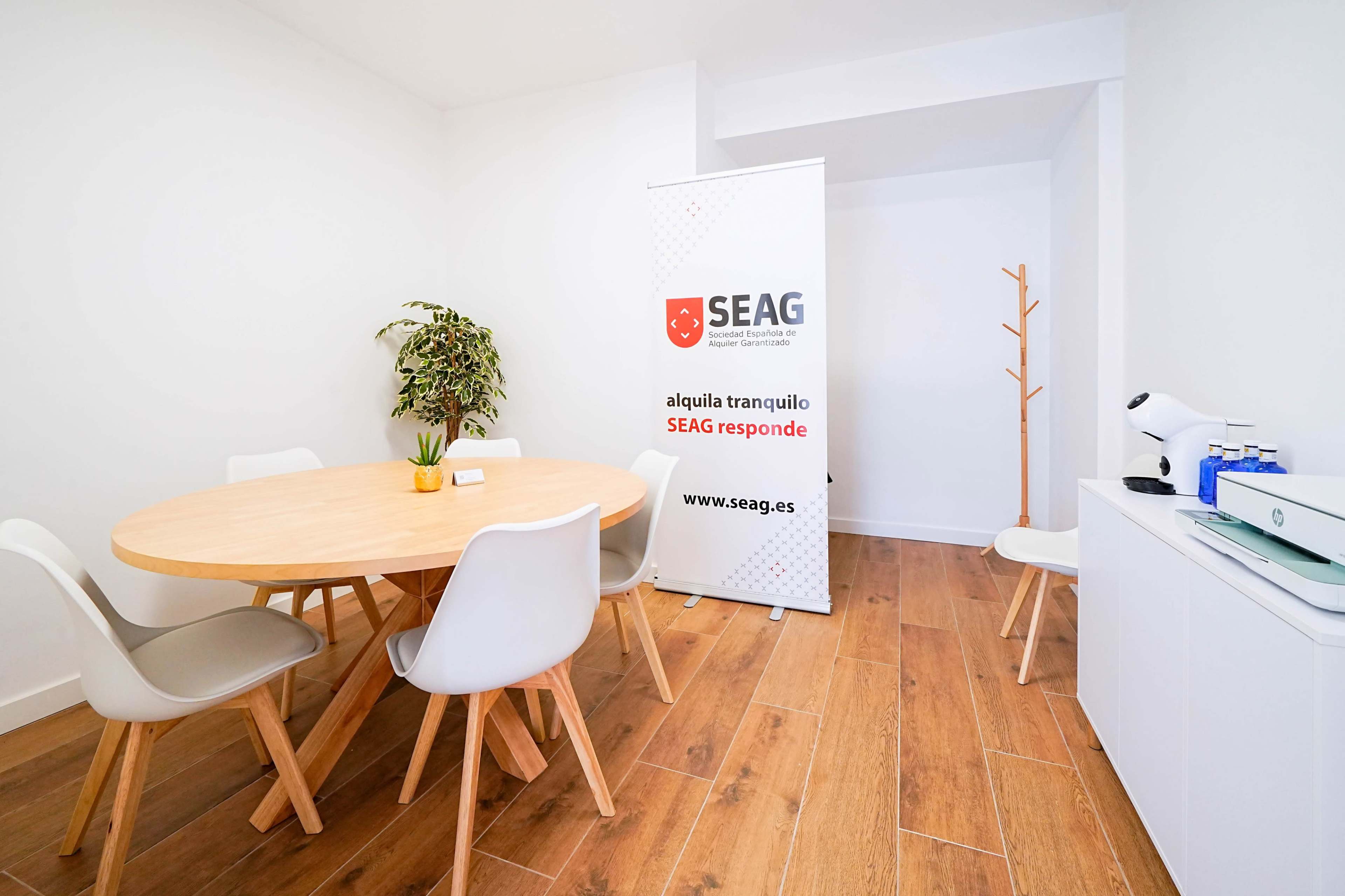 A small, modern meeting room features a round wooden table surrounded by white chairs, with a potted plant, a promotional banner for SEAG, and a coat rack in the corner.