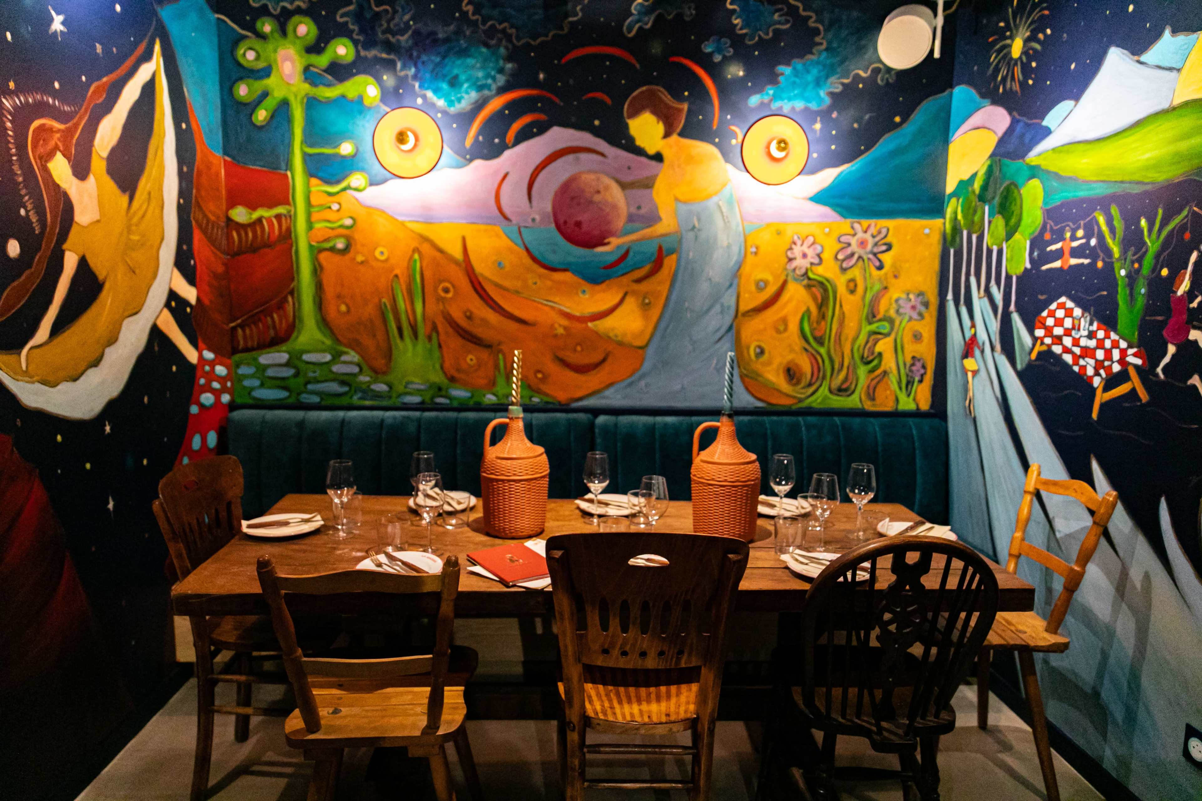 A dining table set for a meal in front of a brightly painted mural featuring colorful scenes and figures.