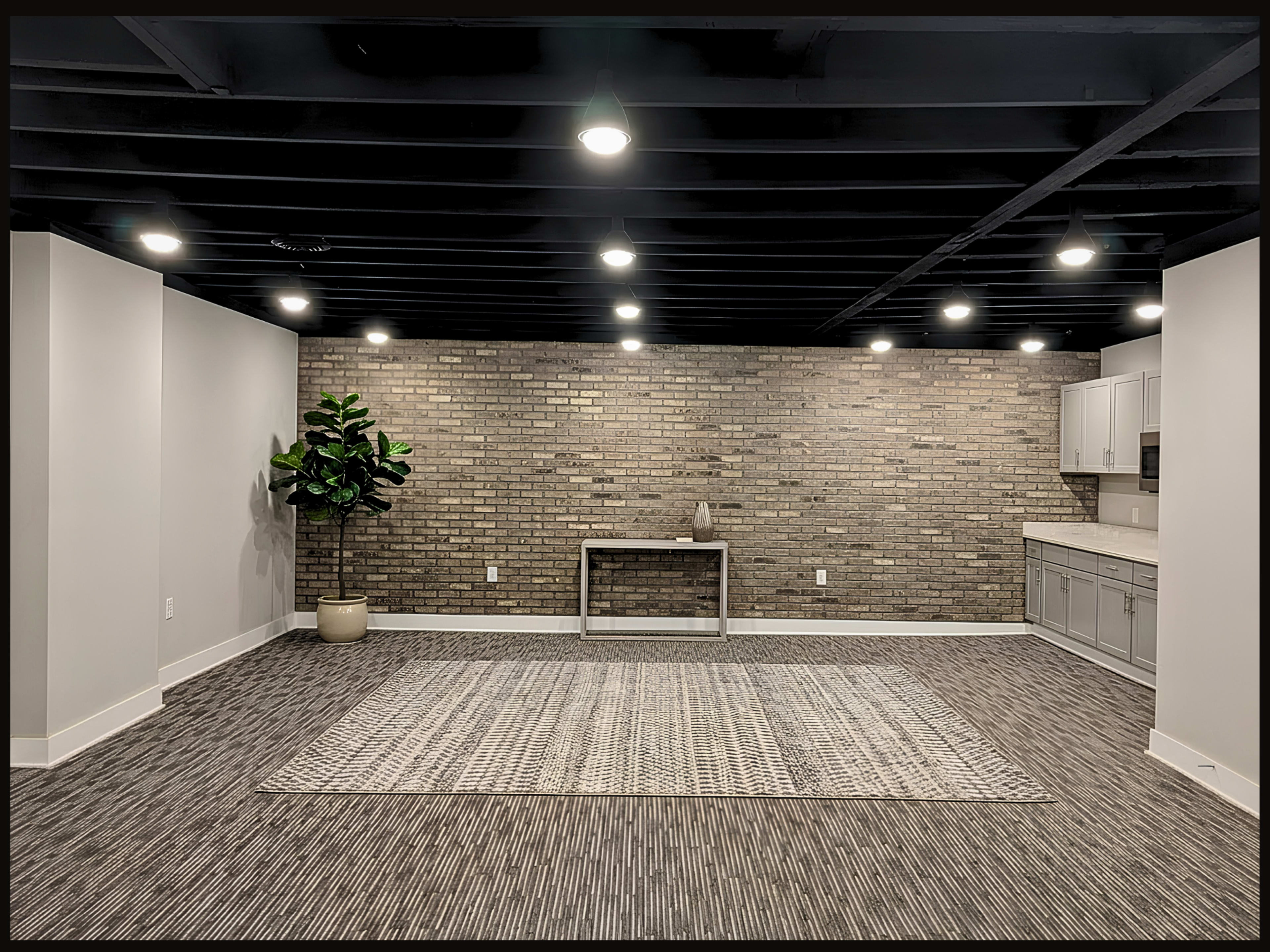 The image shows a spacious, minimally furnished room with a brick accent wall, a table in the center, and a small plant in the corner.