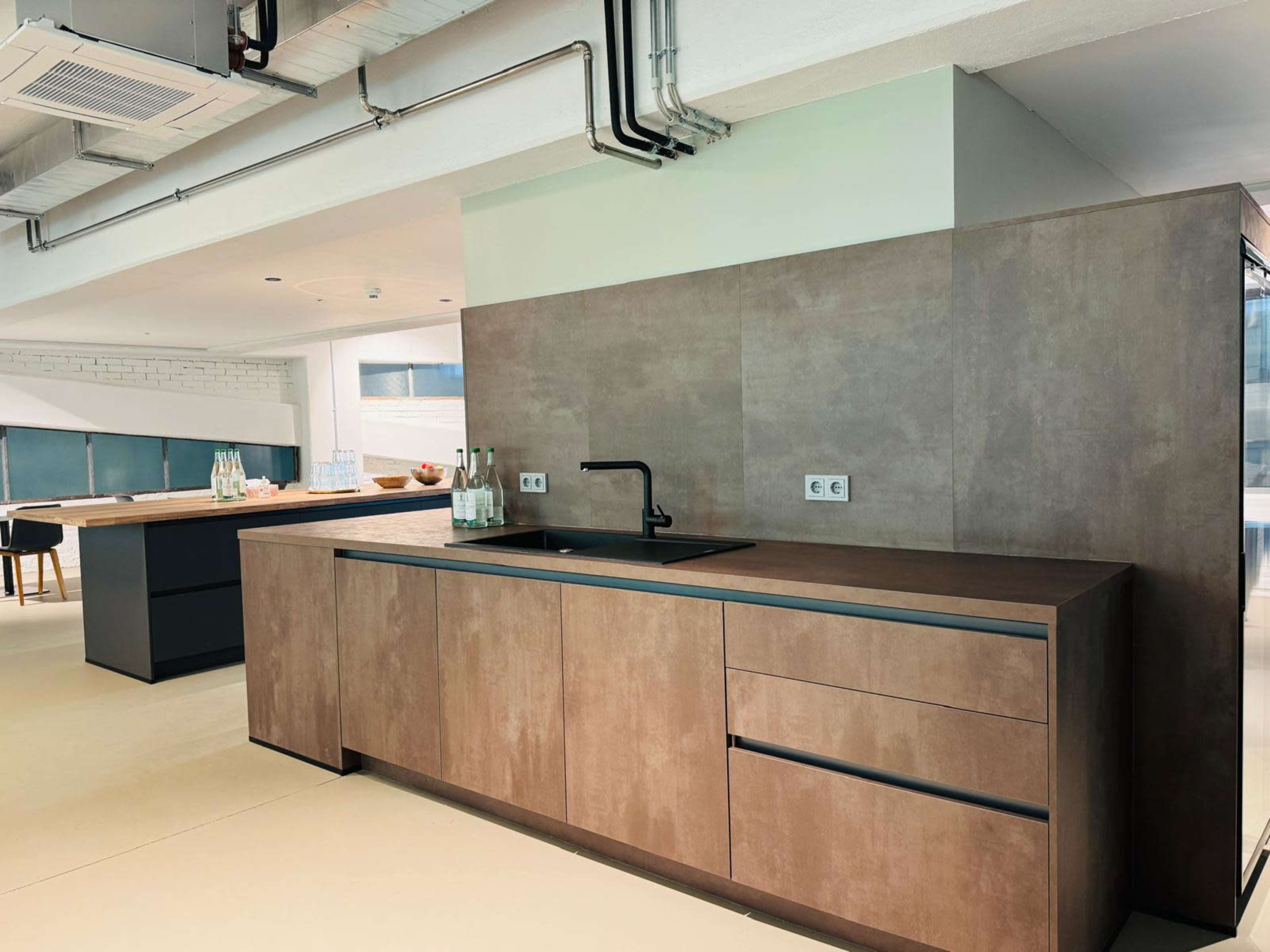 A modern kitchen with a minimalist design, featuring a dark countertop, a black faucet, and simple cabinetry against a light-colored floor.