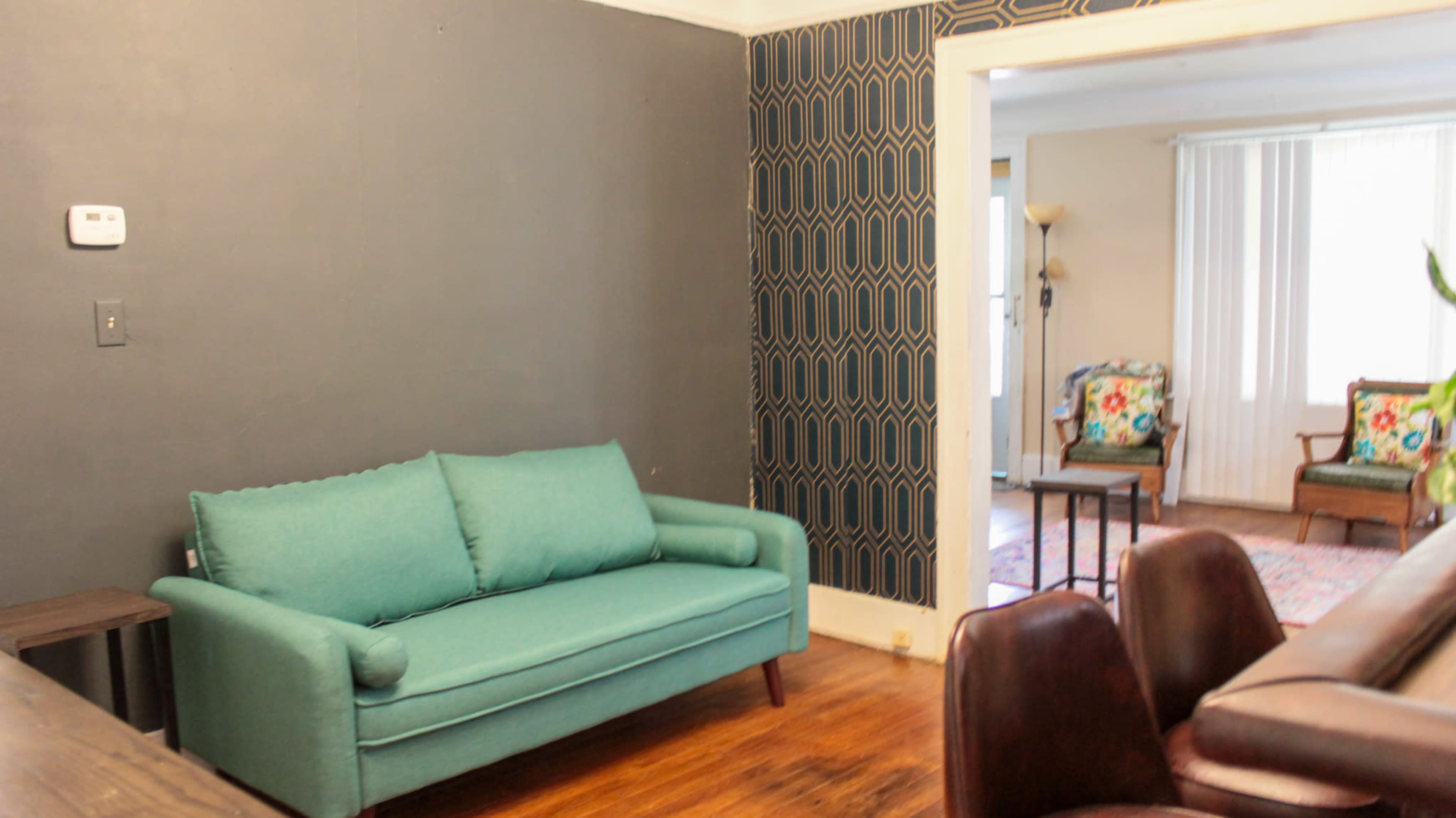 A living room with a teal couch against a gray wall, complemented by patterned wallpaper and visible furniture in an adjacent room.