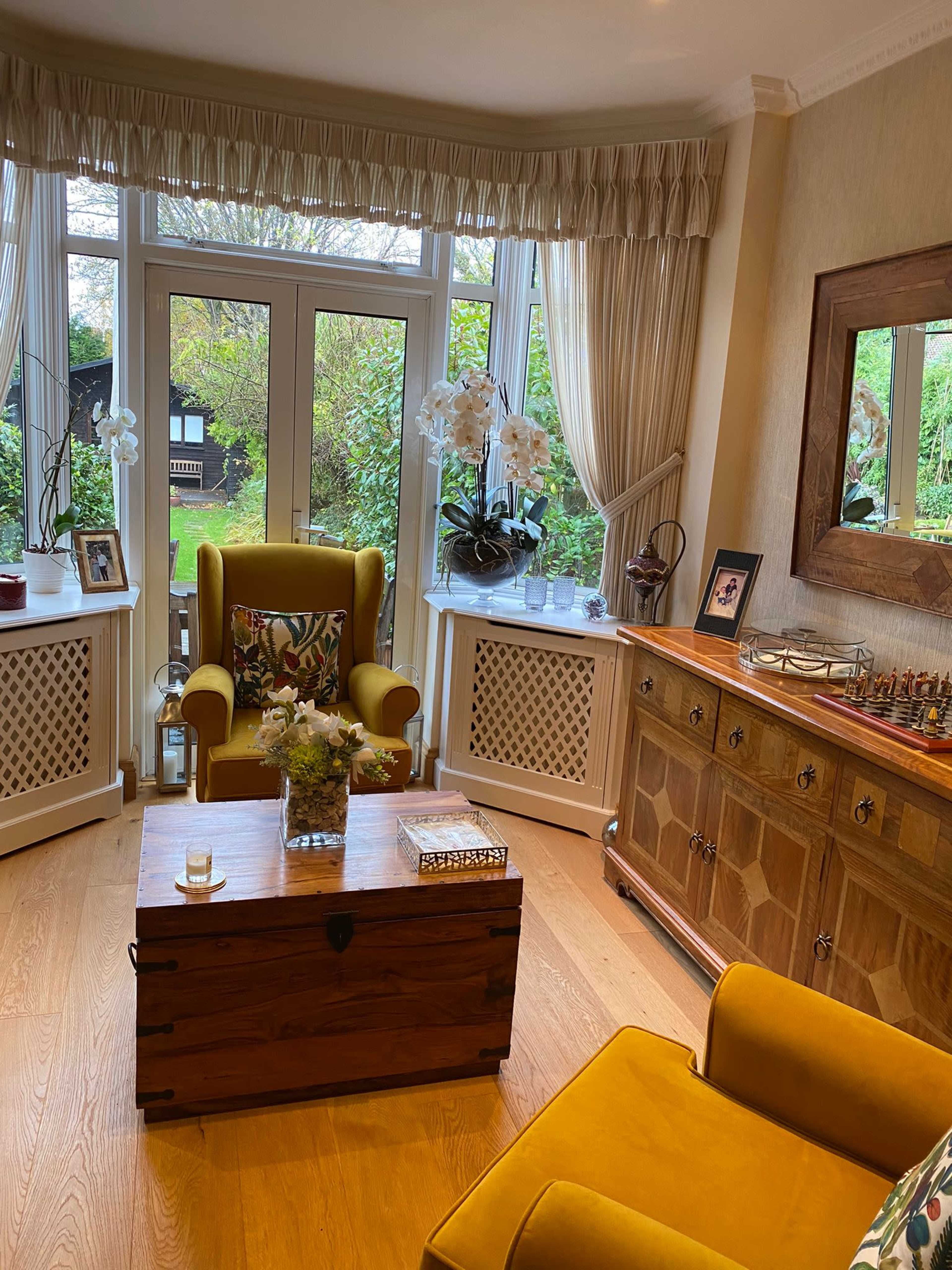 The living room features a yellow armchair, a wooden chest coffee table, and large windows overlooking a garden.
