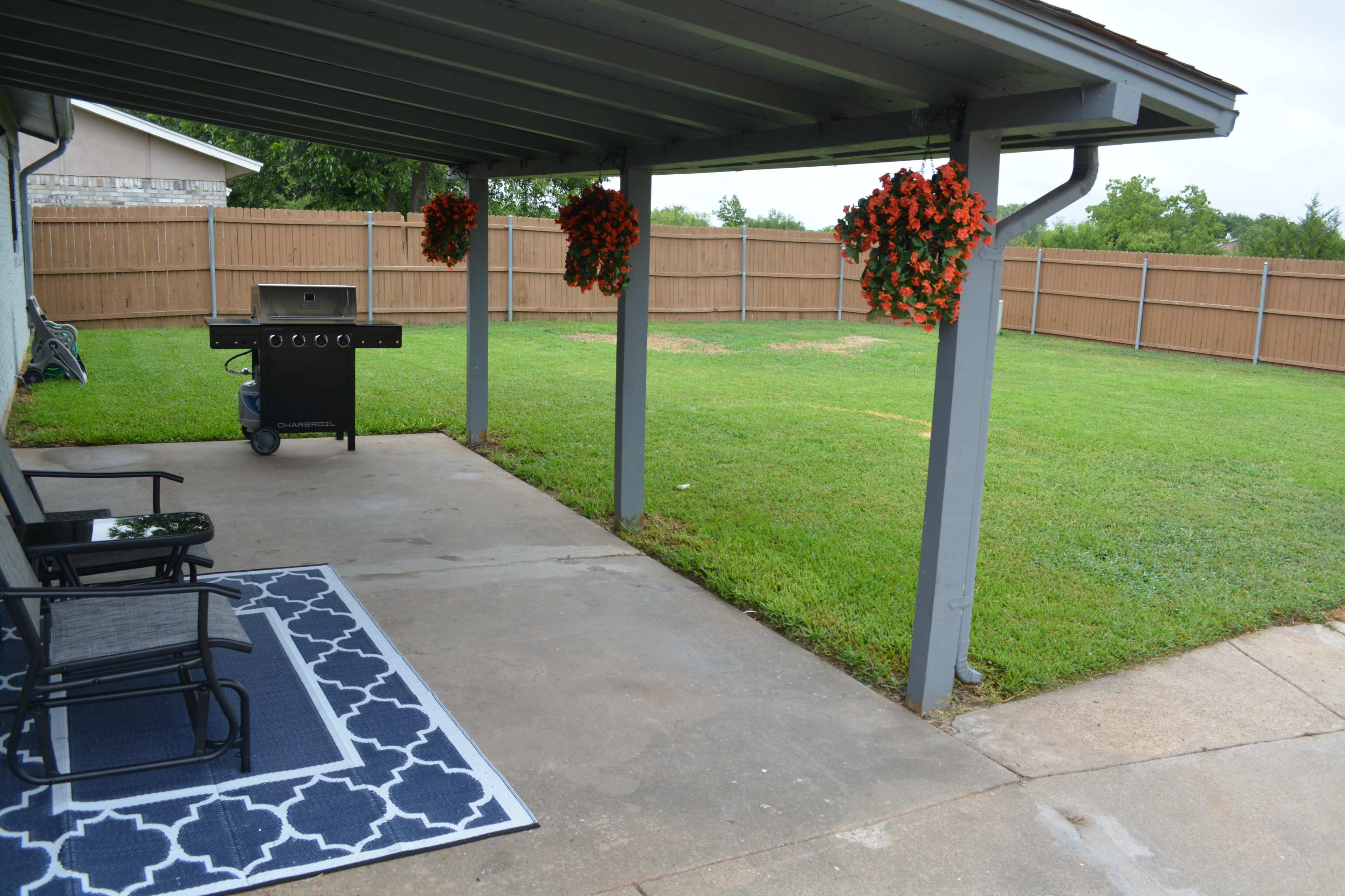 The image shows a covered patio area with a barbecue grill, two black chairs, and hanging flower pots, overlooking a manicured grassy yard enclosed by a wooden fence.