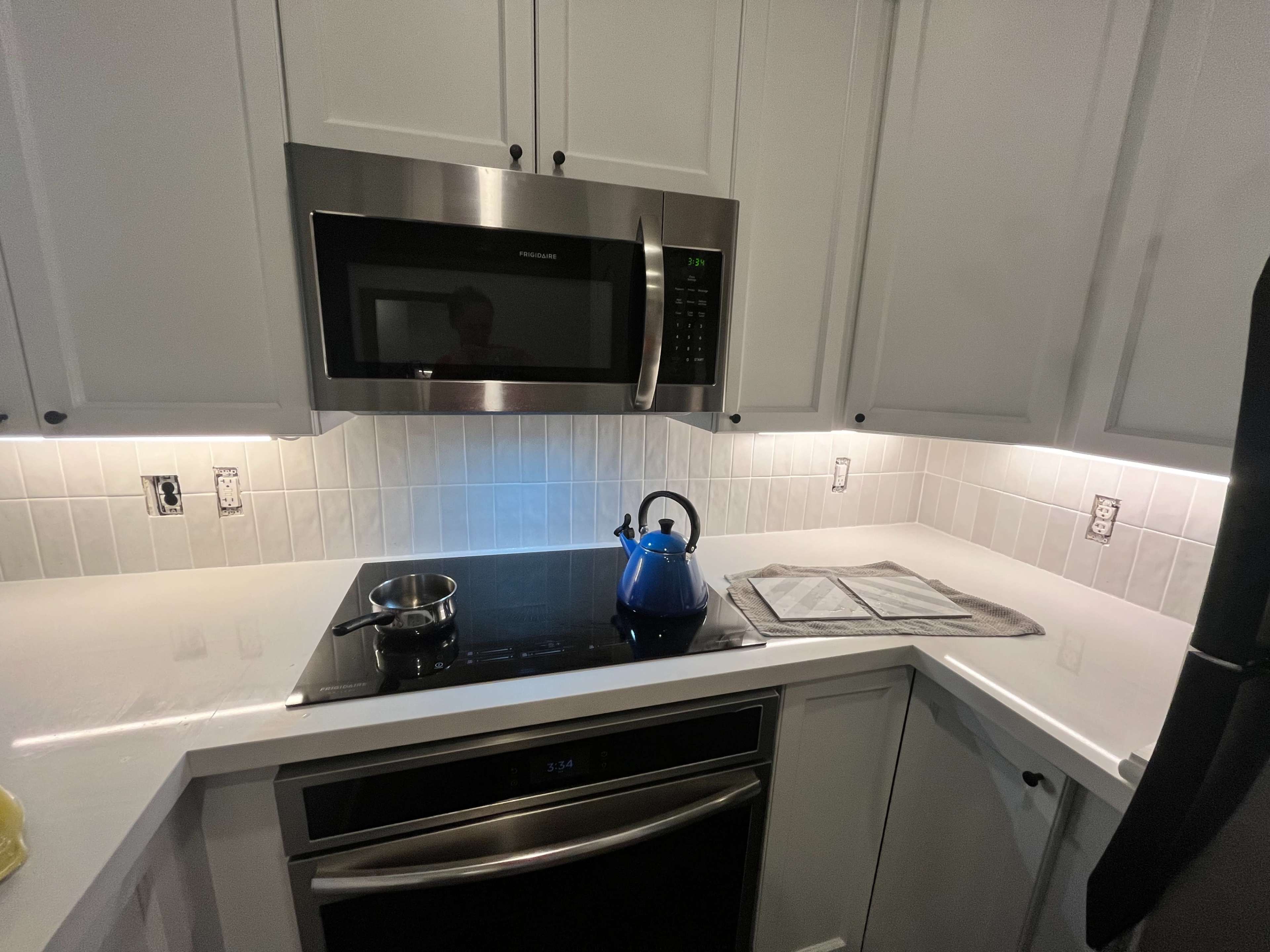 A modern kitchen features a stainless steel microwave above a black induction cooktop with a kettle and a folded towel on the countertop.