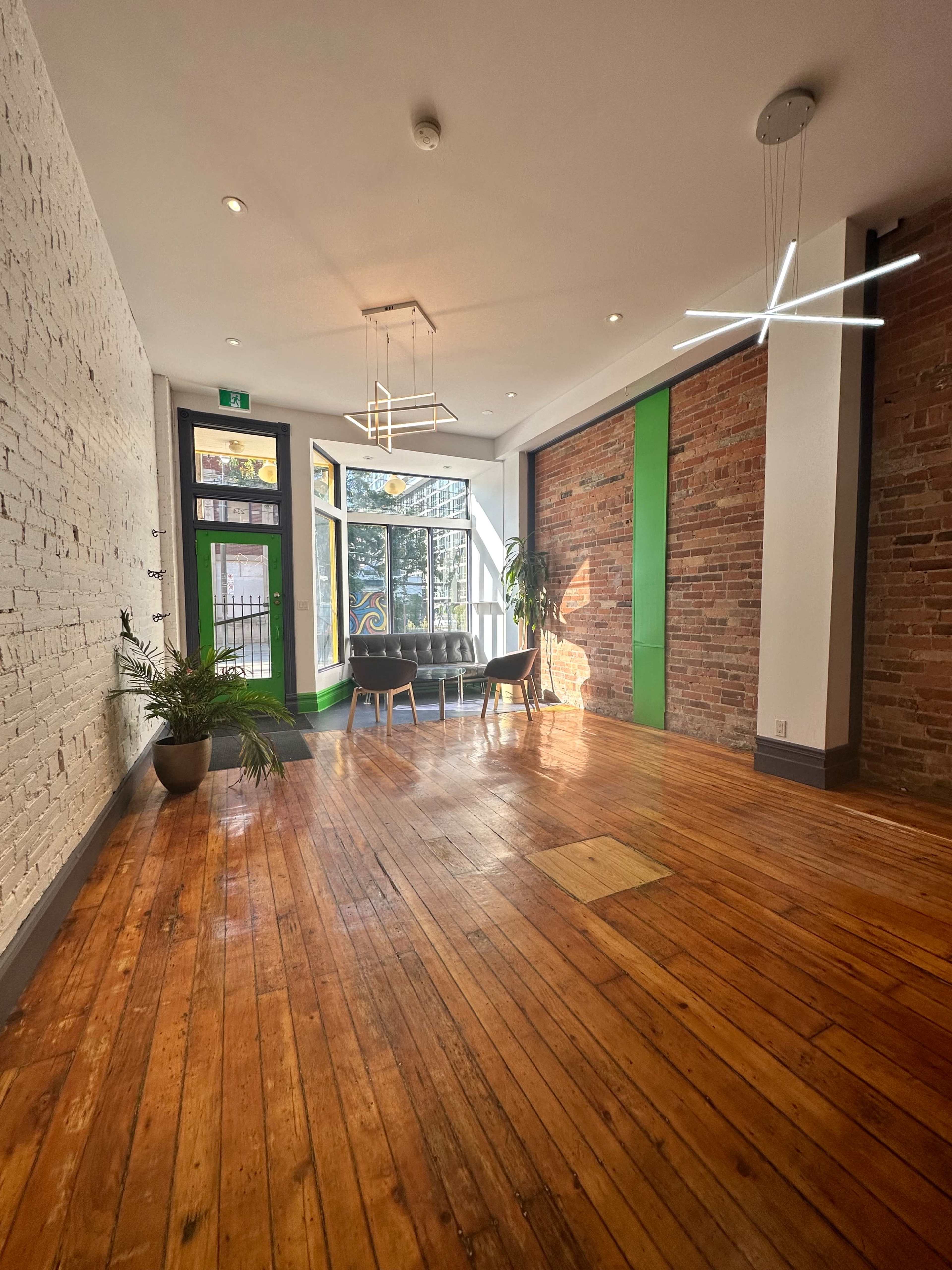 A spacious room with wooden floors, a combination of exposed brick and white walls, modern lighting fixtures, and a seating area near large windows.