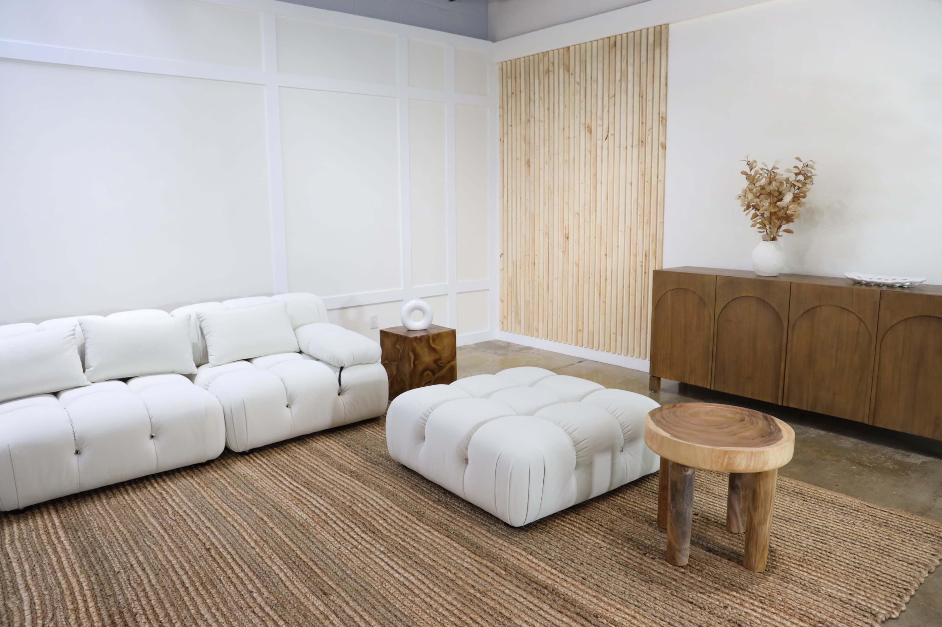 A minimalist living room featuring a white sectional sofa, a tufted ottoman, a wooden side table, and a wooden cabinet against a textured wall.