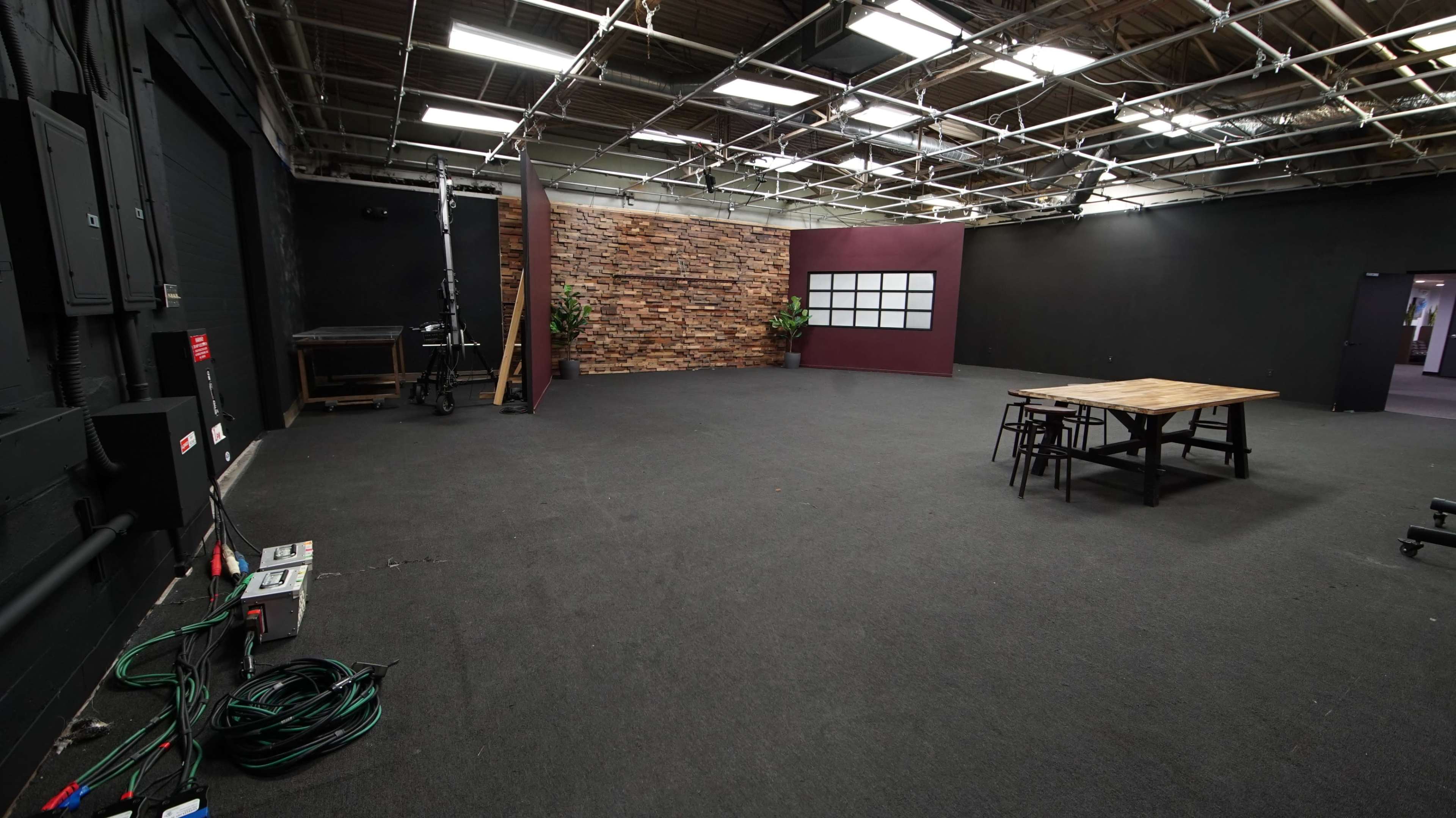 A spacious, empty studio with a textured stone wall, a large table, and a few chairs arranged on a carpeted floor.