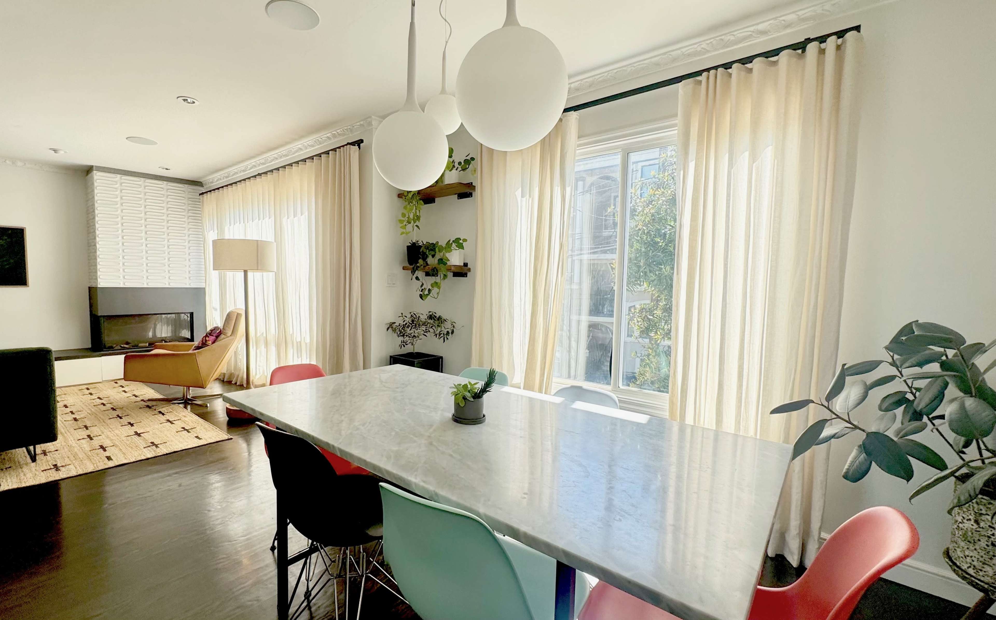 A dining area featuring a marble table surrounded by colorful chairs, with large windows dressed in sheer curtains and plants on the windowsill.