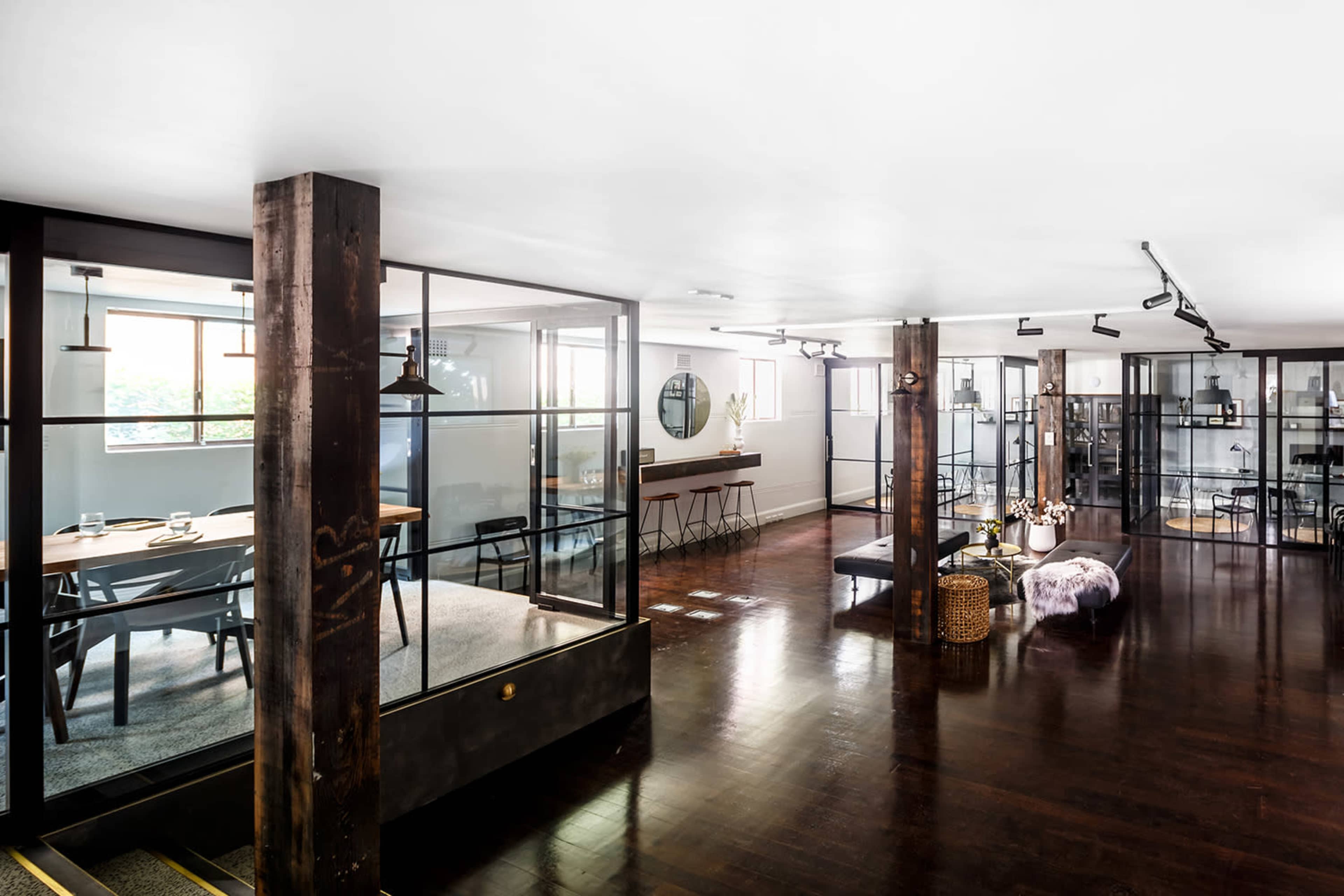 The image shows a modern office space featuring glass-enclosed meeting rooms, wooden beams, and various seating arrangements.