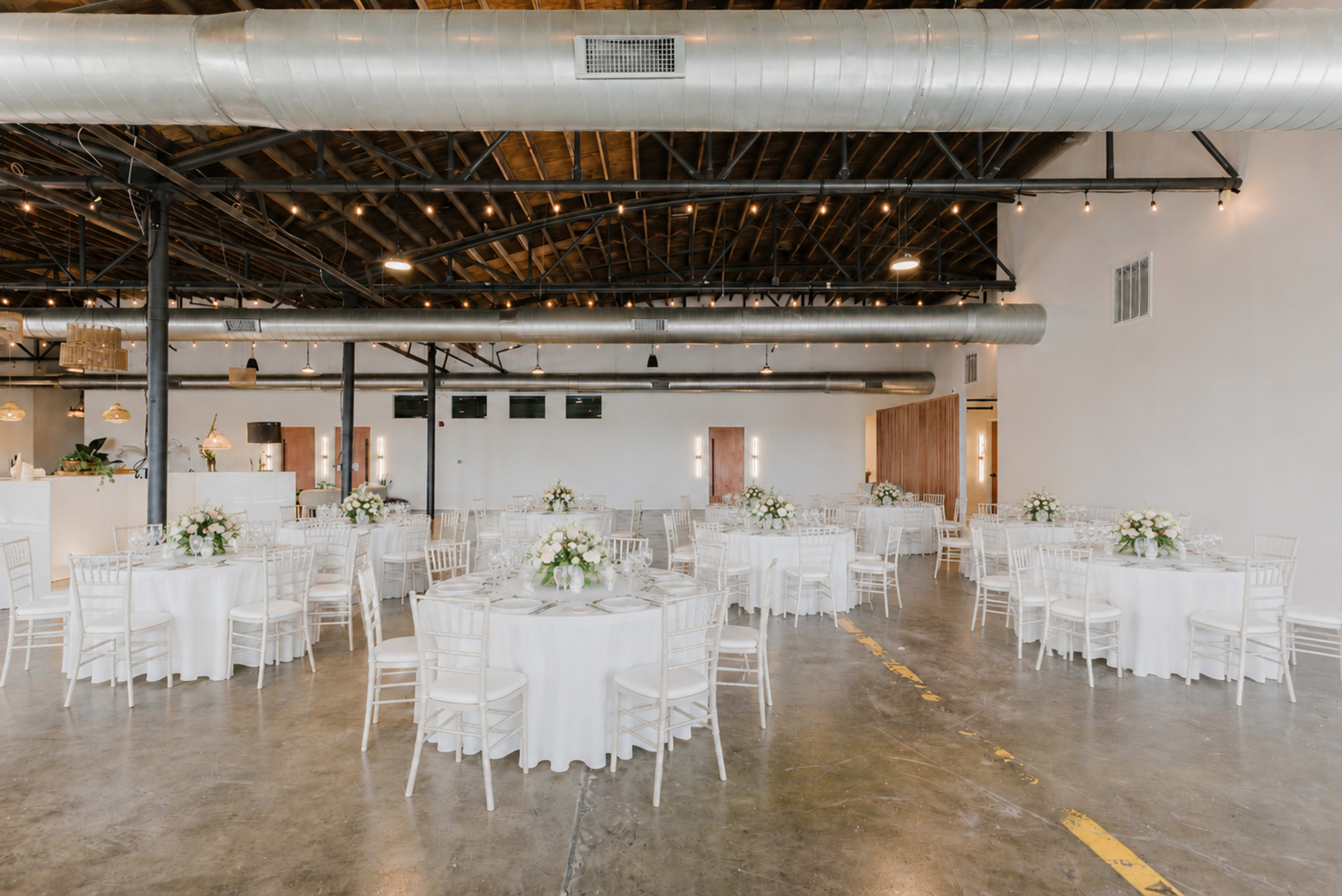 A spacious event venue features circular tables draped in white tablecloths, each adorned with floral centerpieces, set beneath exposed beams and metal ductwork.