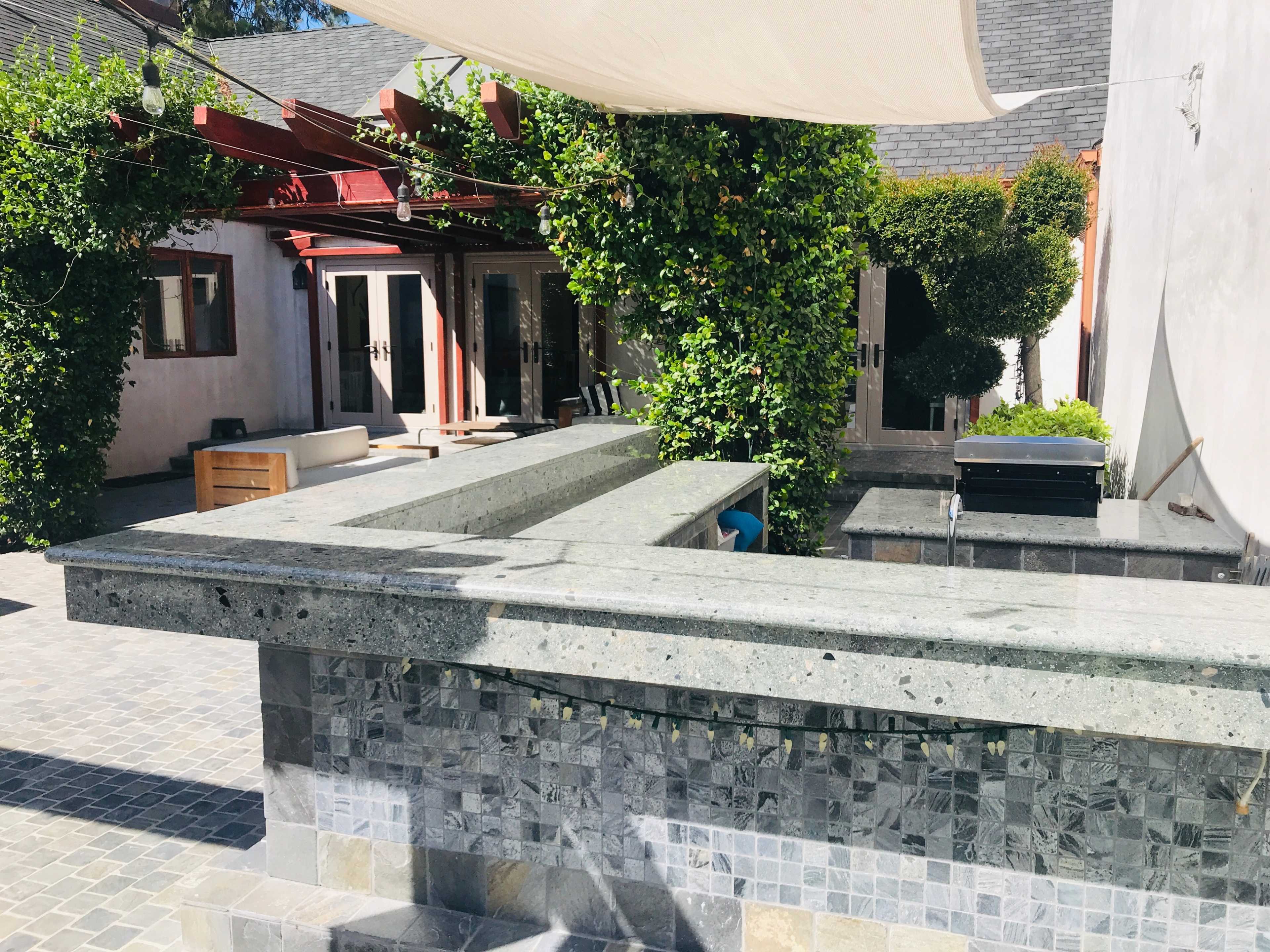 A backyard patio area featuring a stone countertop and a shaded seating area surrounded by greenery and a house in the background.