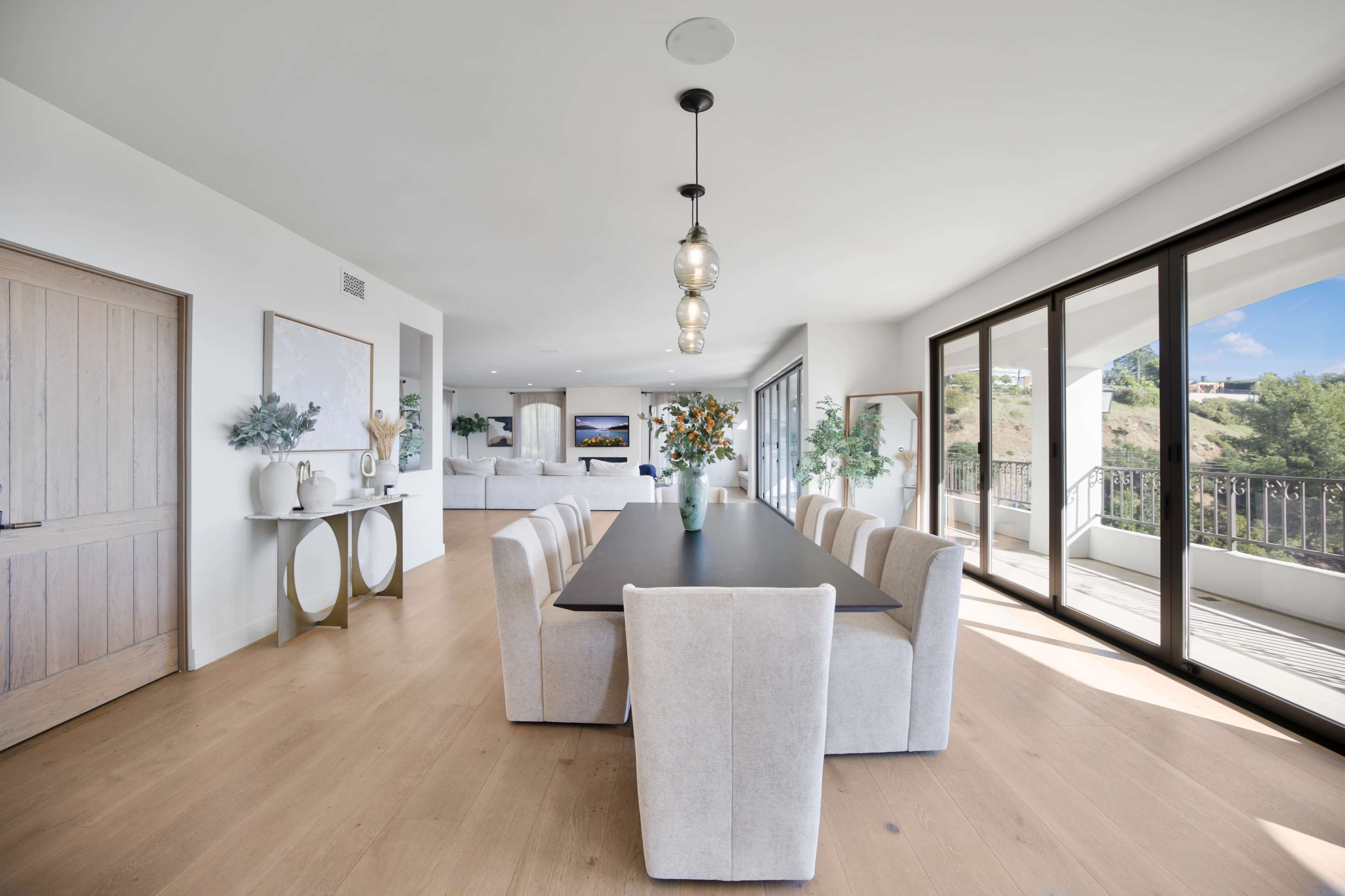 The image shows a spacious dining area with a long table surrounded by upholstered chairs, large windows, and a view of an outdoor patio.
