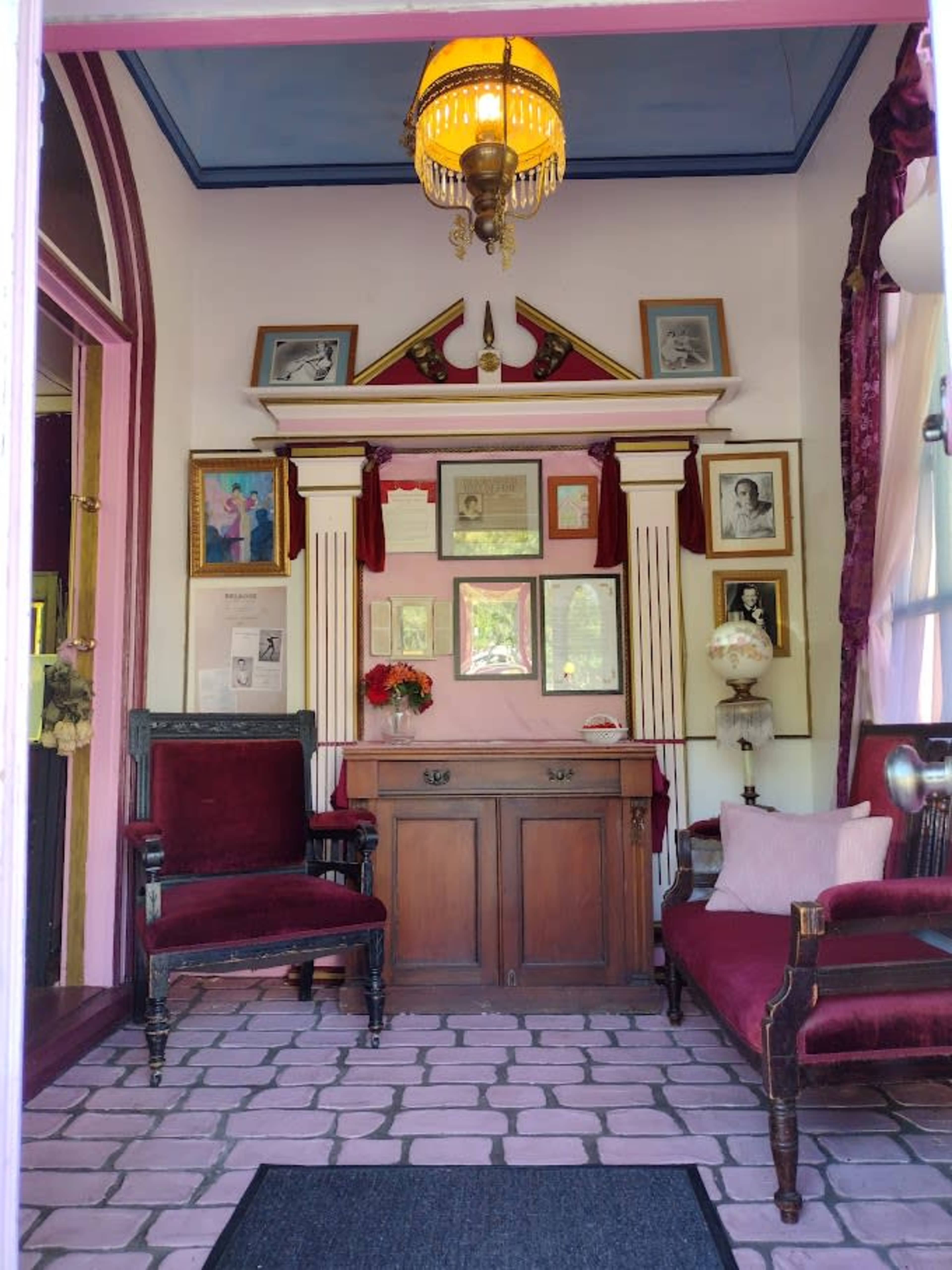 The image shows a small, elegantly furnished room with two chairs, a vintage lamp hanging from the ceiling, and walls adorned with framed photographs and articles.