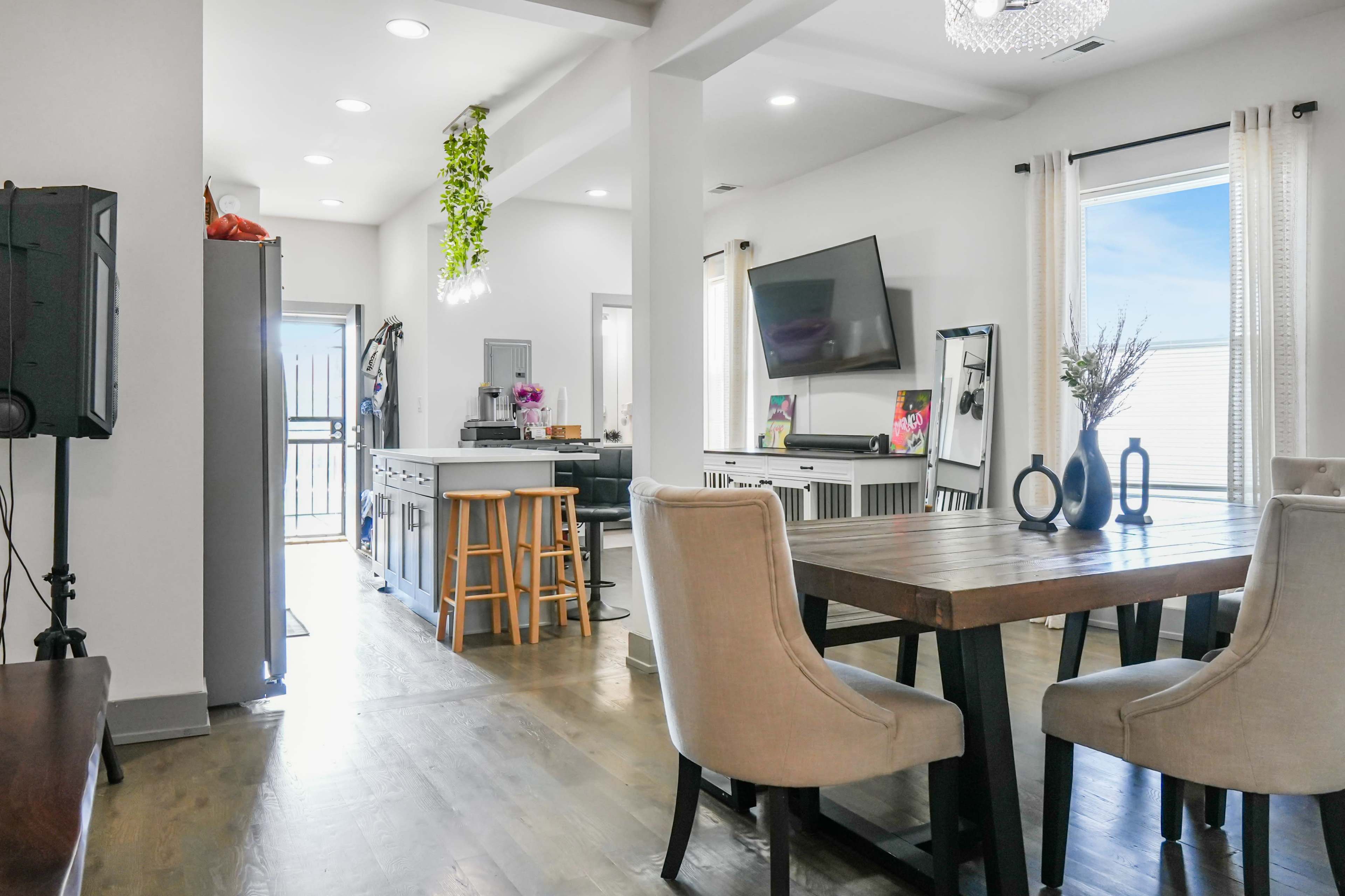 The image shows an open-concept living space featuring a dining area with a wooden table and upholstered chairs, leading to a kitchen with modern appliances and a door to an outdoor space.