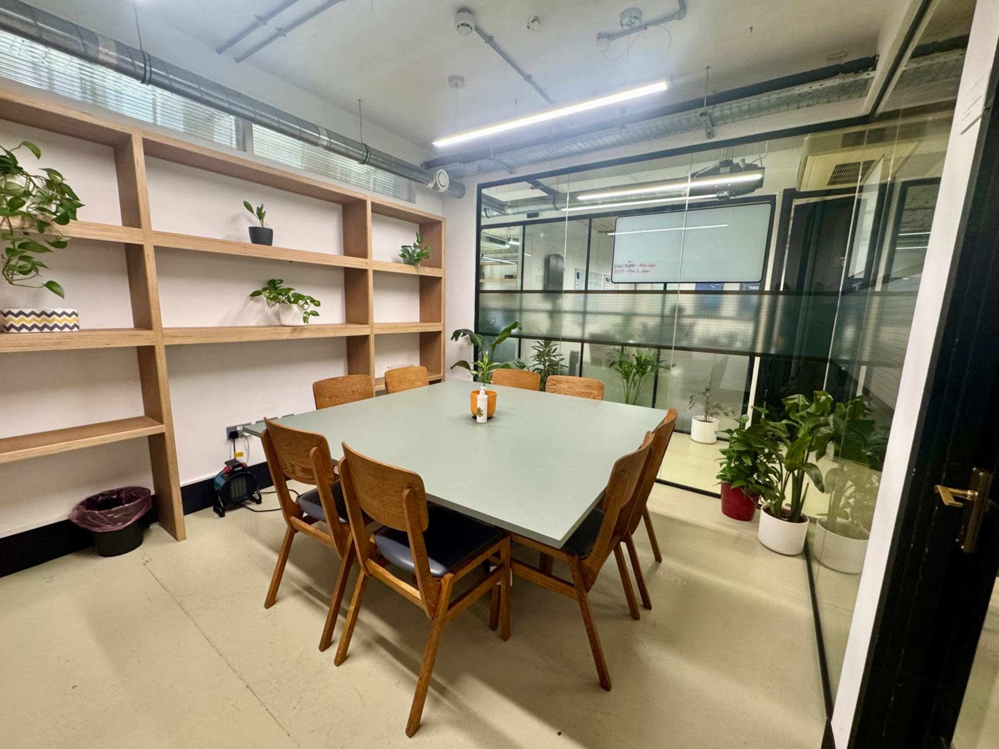 The image shows a modern meeting room with a large table surrounded by wooden chairs, and shelves filled with potted plants in the background.