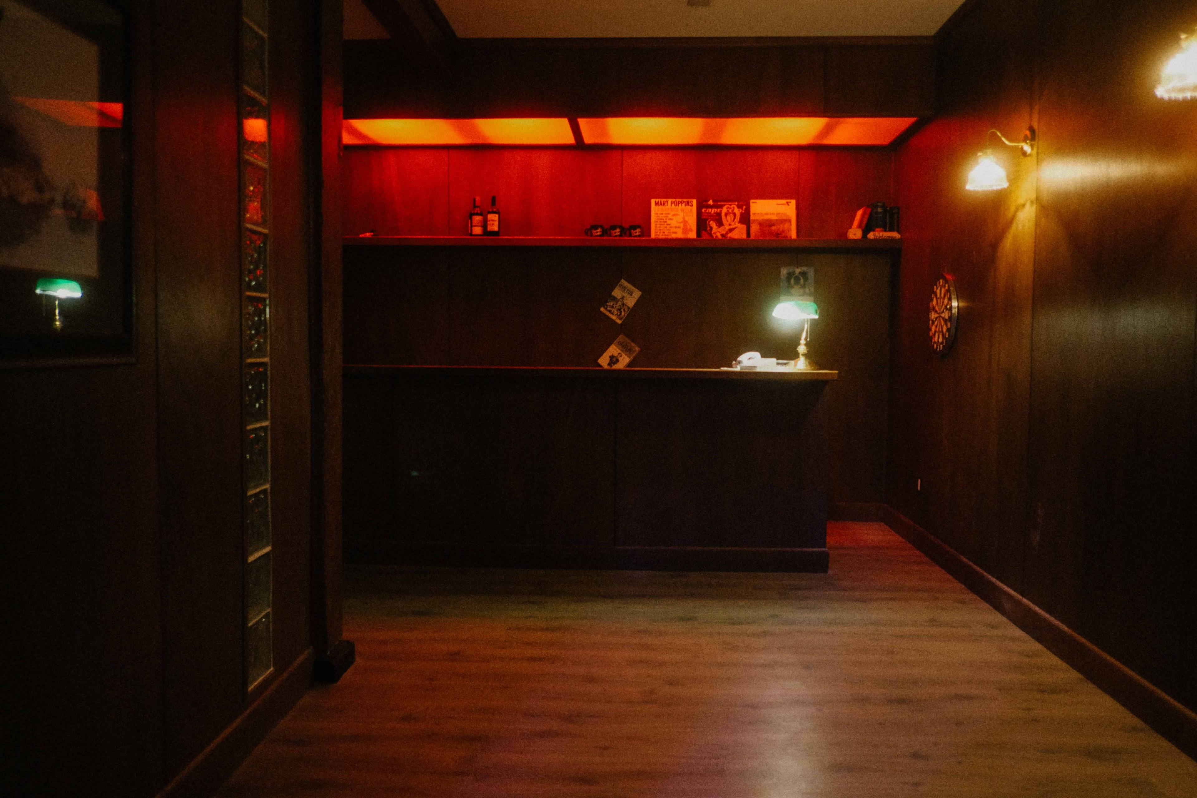 The image shows a dimly lit interior of a bar with a wooden counter, accented by red lighting and a dartboard on the wall.