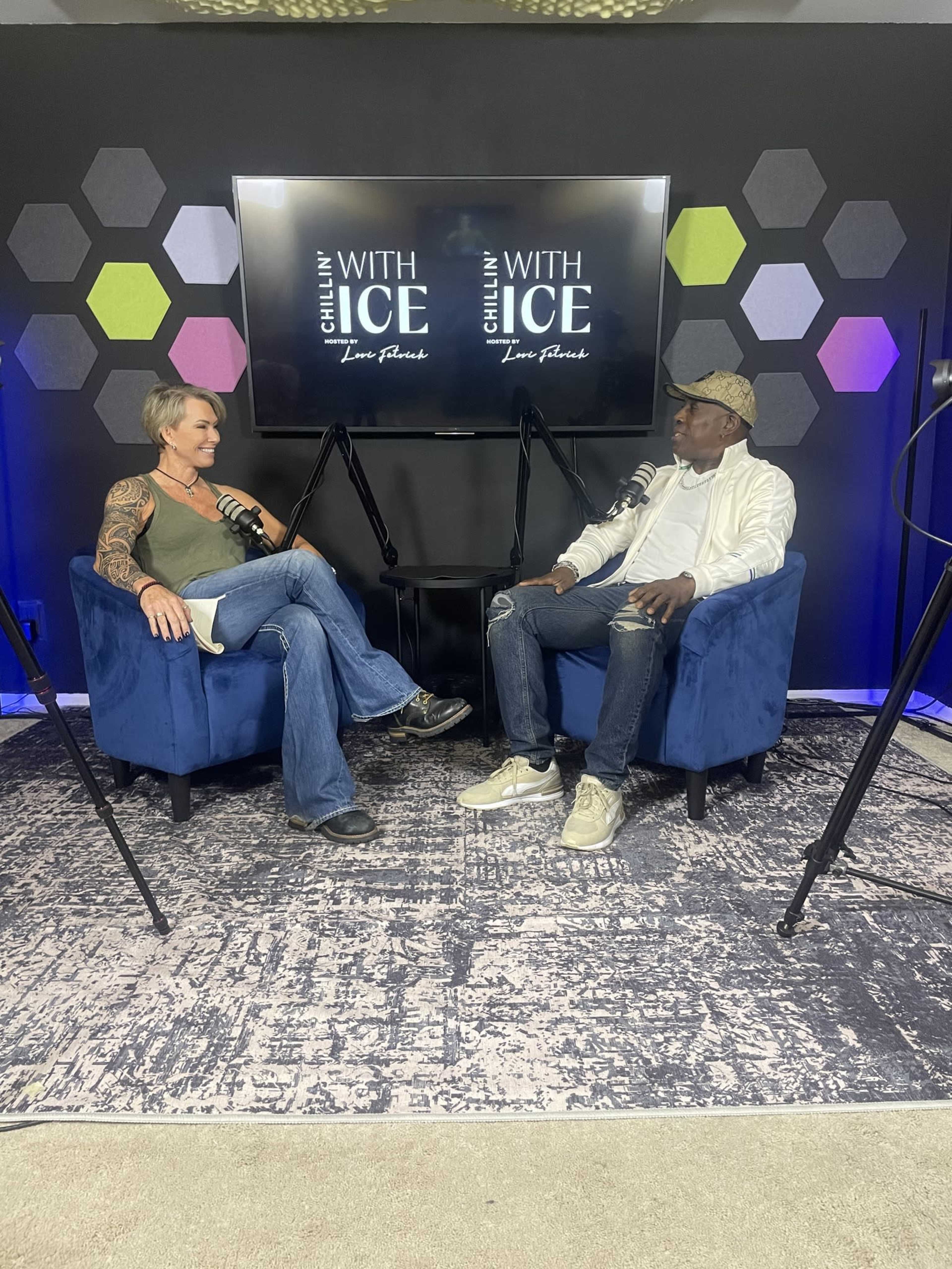 A woman and a man sit facing each other in blue chairs, engaged in conversation, with microphones in front of them, in a studio setting featuring geometric wall designs.
