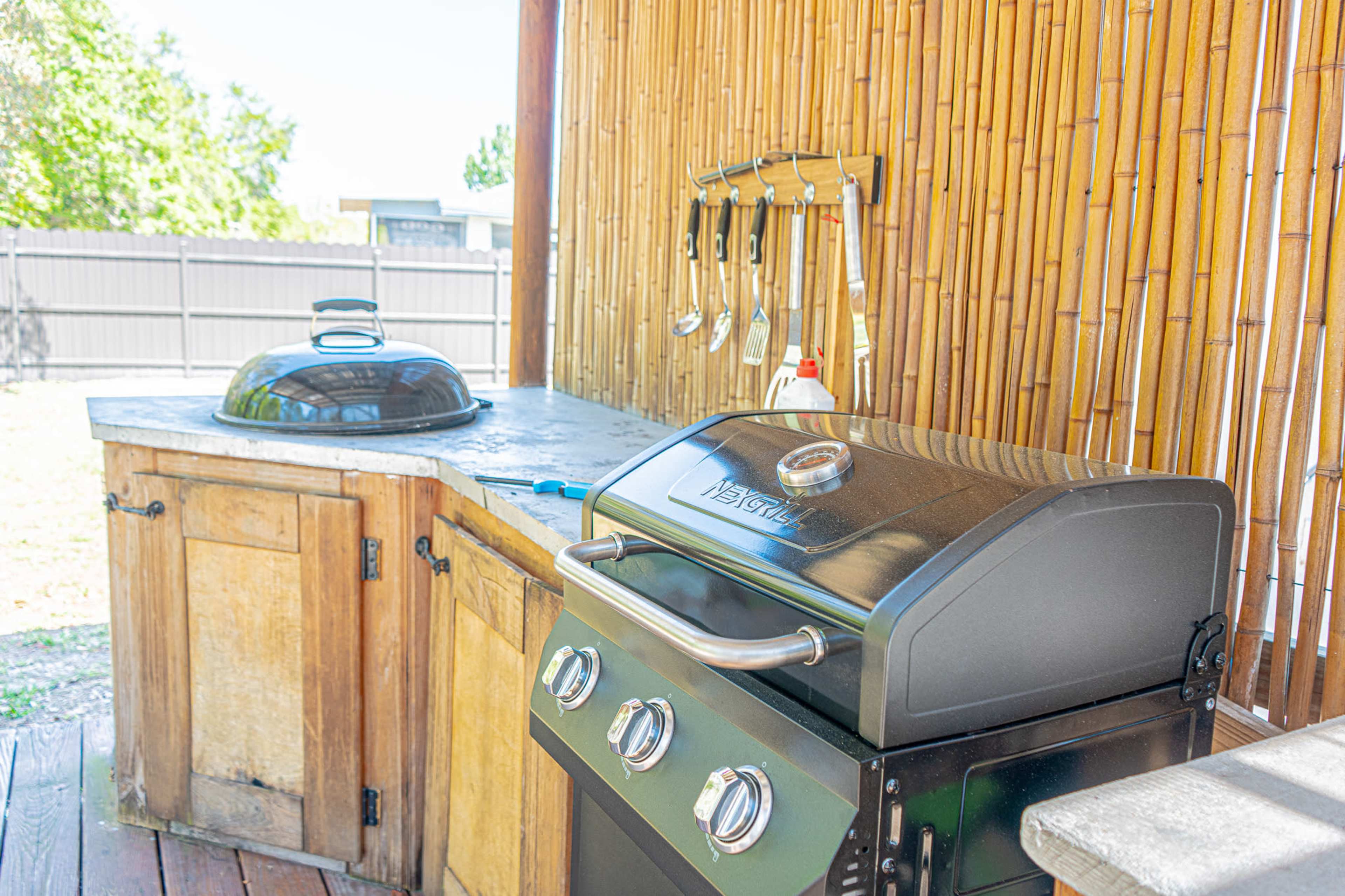 The image shows an outdoor cooking area featuring a gas grill, a covered pot, and various utensils hanging against a bamboo backdrop.
