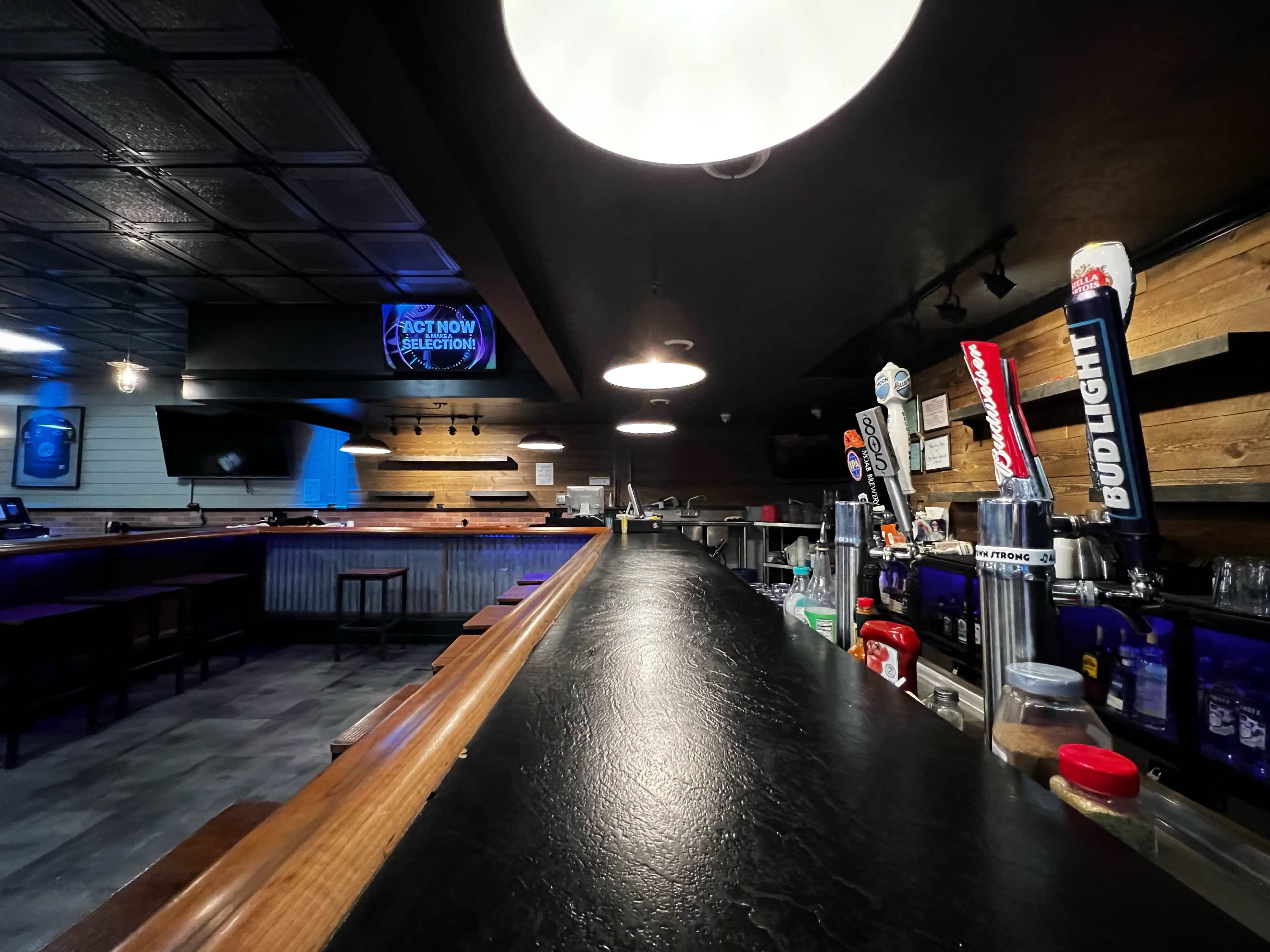 The interior of a bar with a long, dark countertop, various beer taps, and a menu sign illuminated in blue light.