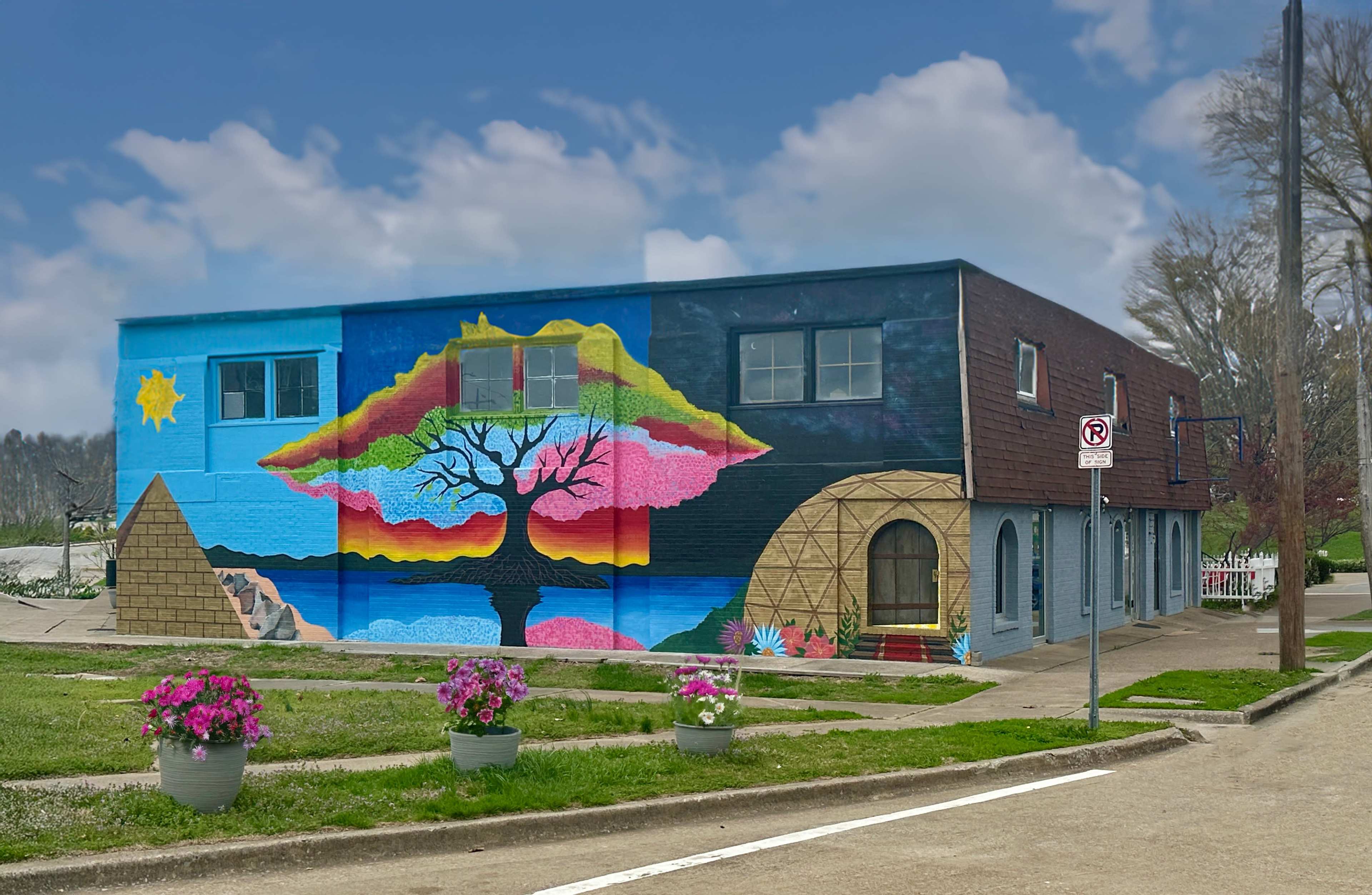 The image shows a colorful mural on the side of a building, featuring a stylized tree, mountains, and a sun, with potted flowers in the foreground.